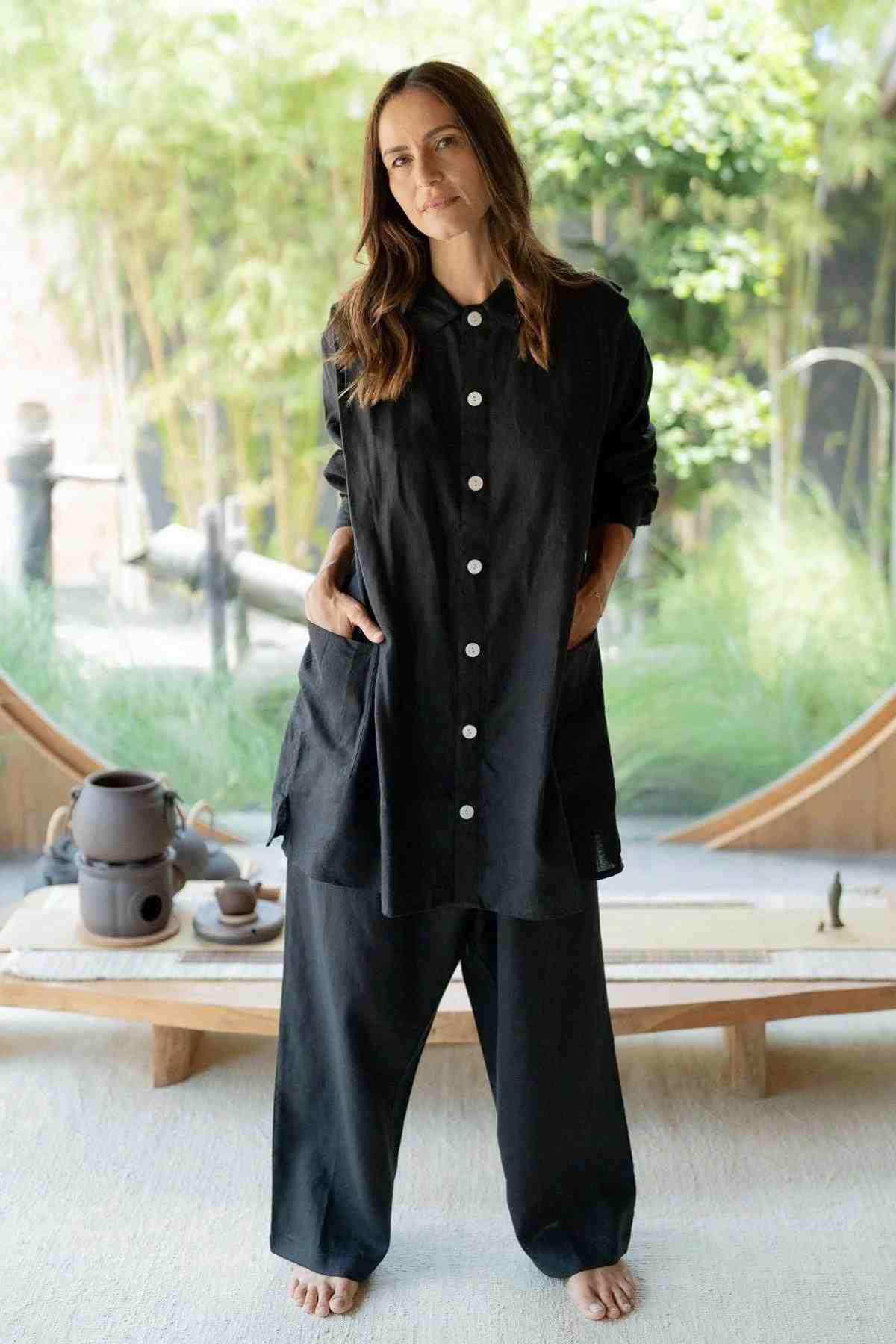 A woman stands barefoot on a light-colored floor, wearing a loose-fitting black outfit with buttons on the top. She has her hands in her pockets and stands in front of a circular window that frames a lush green garden. An eclectic fashion statement, her ensemble is complete with the Tender Jacket (100% Linen) by Myrah Penaloza. A low wooden table with a tea set rests behind her.