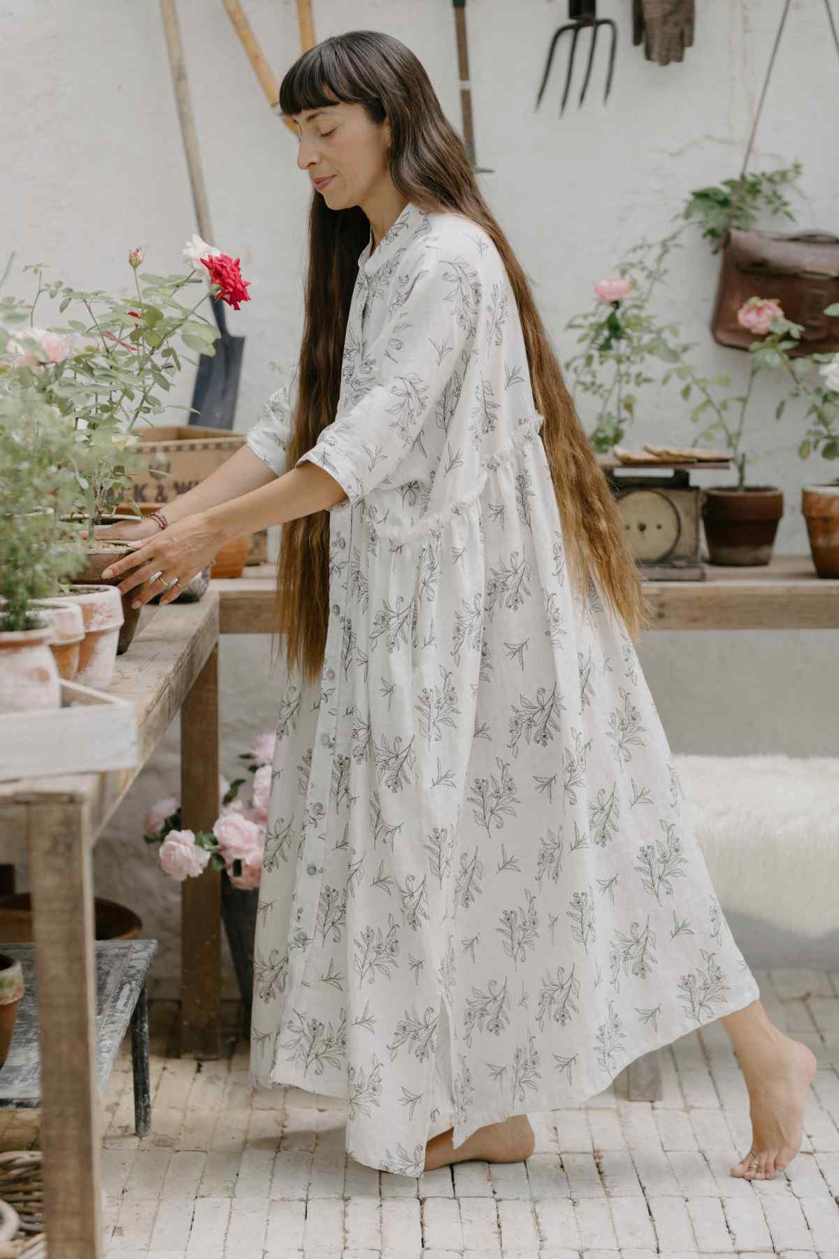 A person with long hair, wearing a flowing Bona Dress from the Cha Dao Tea Collection by Myrah Penaloza, adorned with white floral patterns and crafted from 100% linen, is tending to potted plants on a wooden table in a rustic indoor setting. Various gardening tools and flowers are visible in the background.