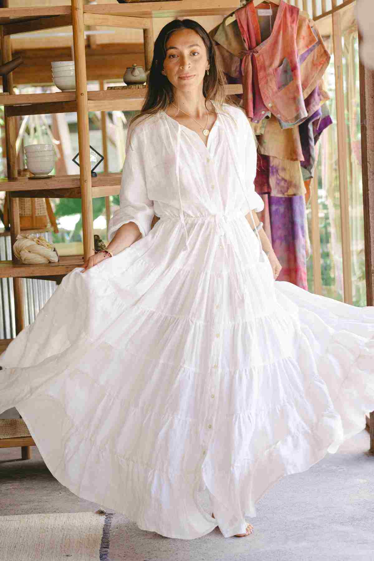 Wearing the La Mexicana Gown by Myrah Penaloza, a person stands indoors. The gown features an adjustable waist and long sleeves with a tiered skirt that cascades elegantly. In the background, colorful textiles add a vibrant touch to the scene, while they stand before a wooden shelf adorned with various objects.