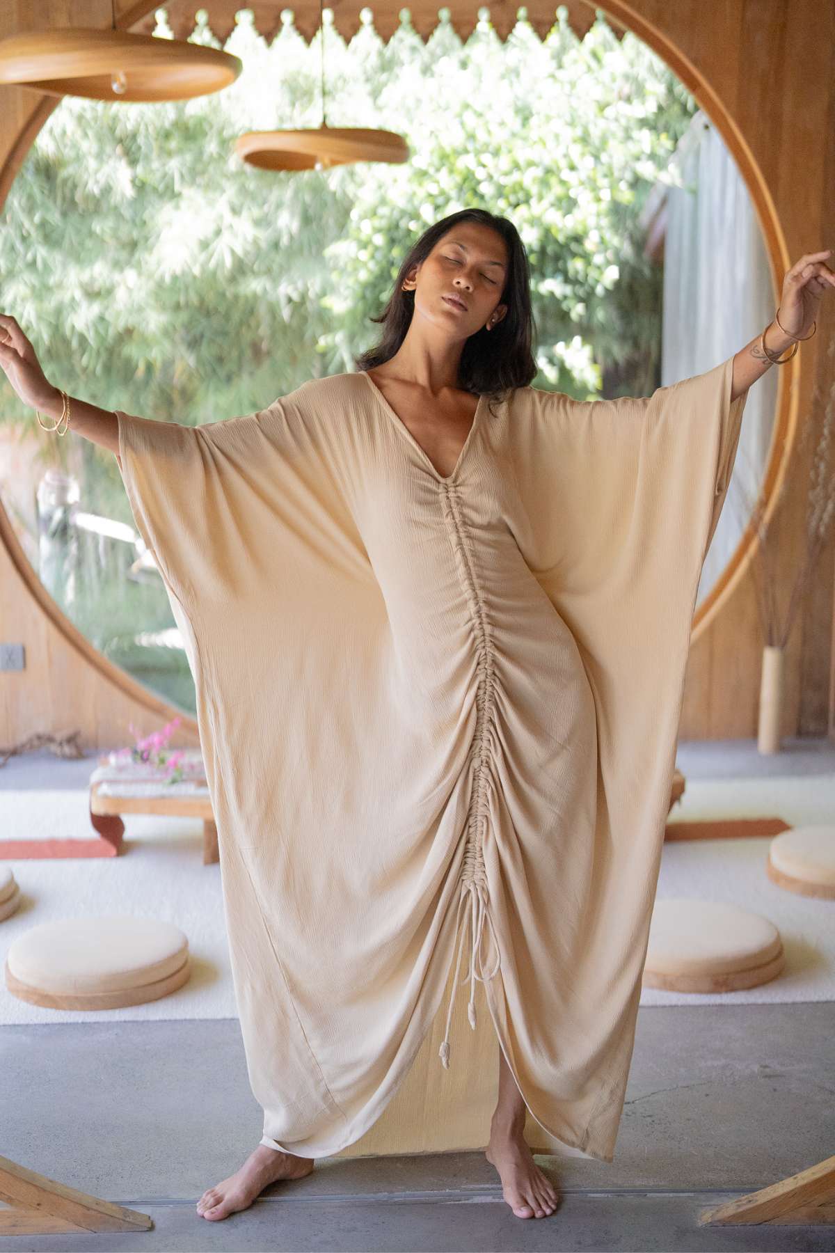 A woman elegantly poses barefoot inside a serene wooden setting, wearing the flowing Virgo Moon kaftan with long sleeves by Myrah Penaloza. A large circular window offers a view of lush greenery outside, as gentle light streams in, accentuating the exquisite details of her attire.