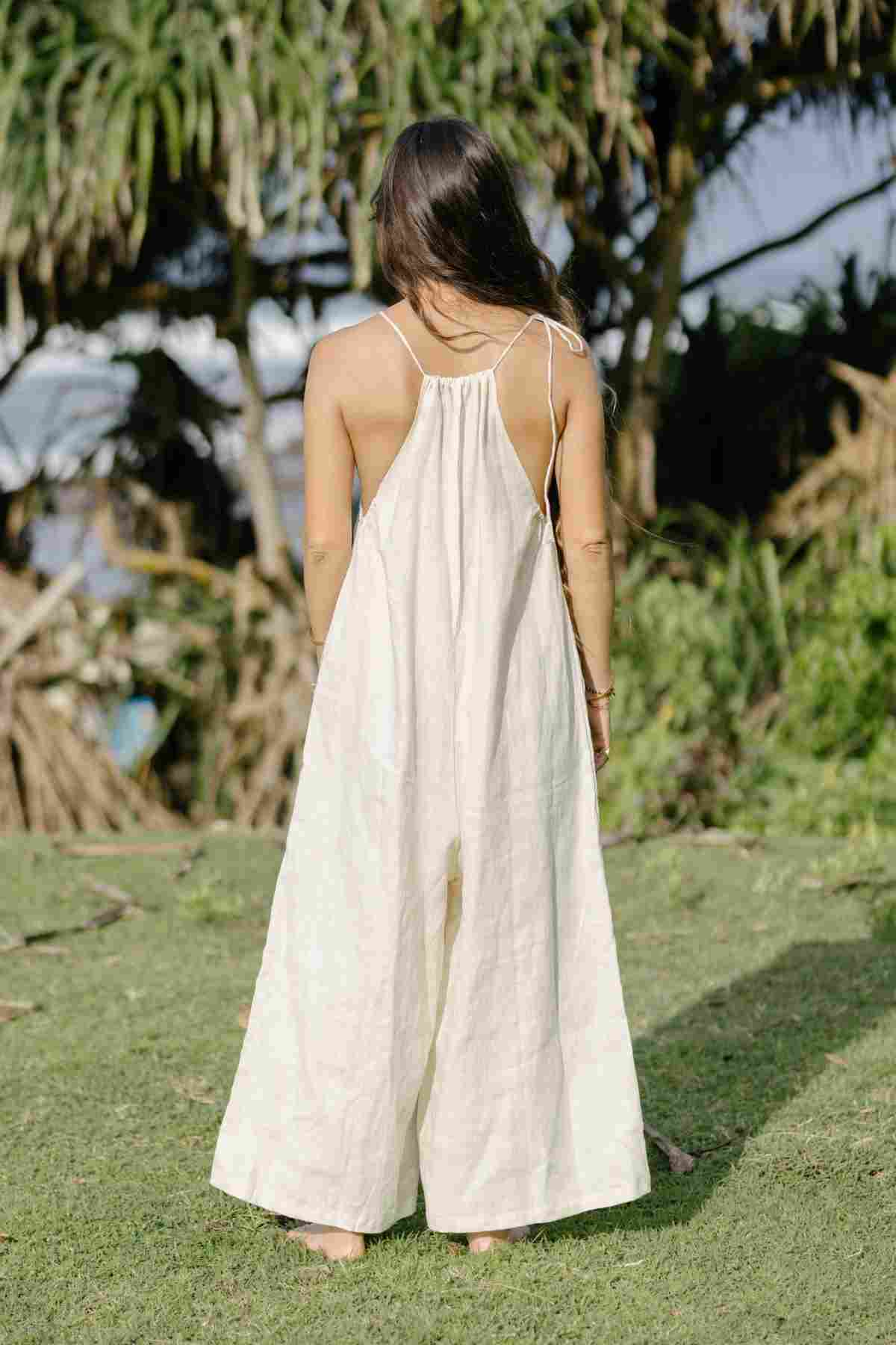 A woman with long brown hair, standing on a grassy field and facing away from the camera, is wearing a long, flowing Linen Vata Playsuit by Myrah Penaloza. The playsuit, made from 100% linen and featuring thin straps, stands out against the backdrop of trees and greenery under a clear sky.