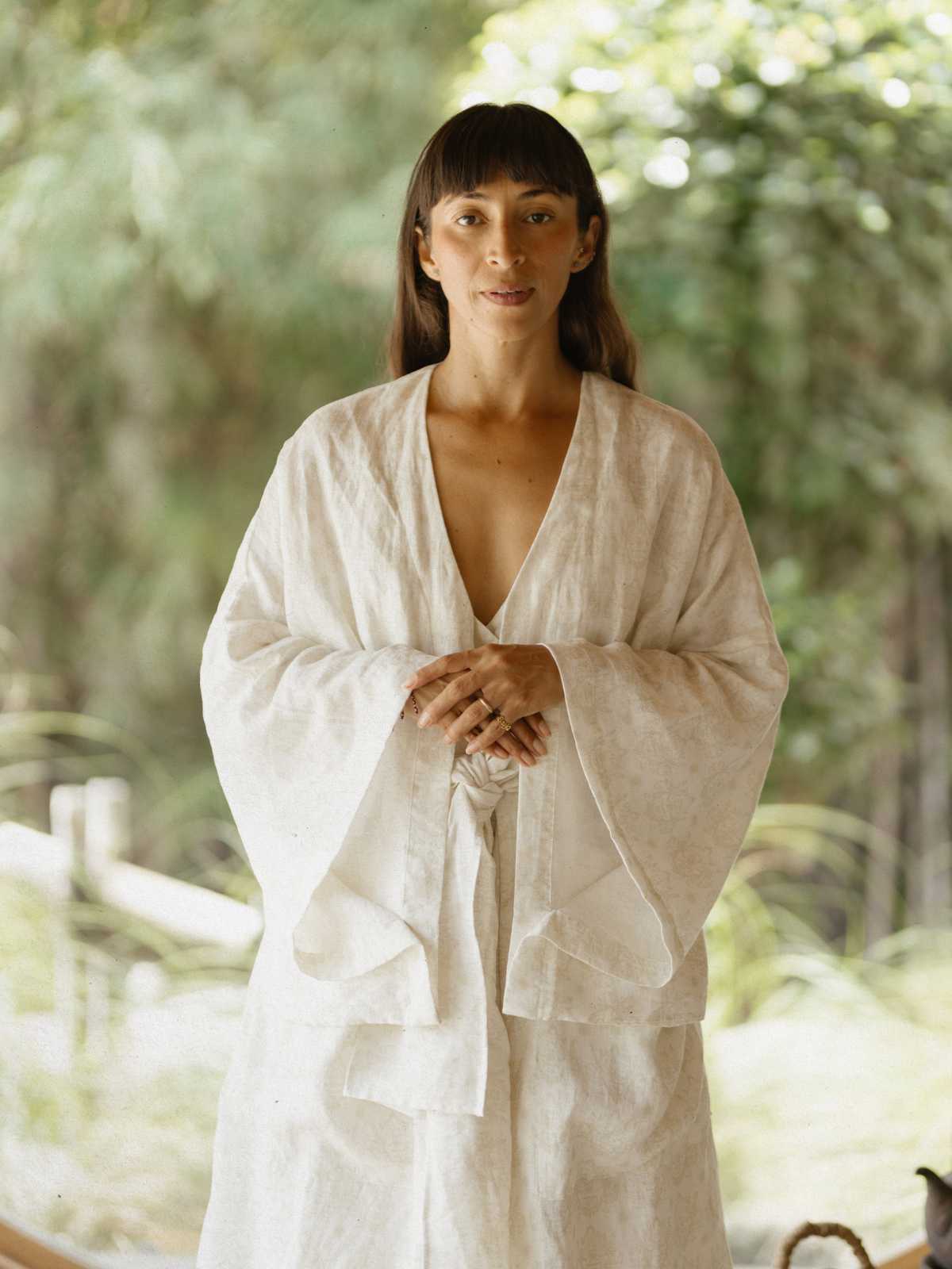 Dressed in a Libra Kimono by Myrah Penaloza, made from 100% linen, a person stands outdoors. Their serene facial expression is framed by their long hair with bangs, while lush green foliage and soft natural light add to the peaceful atmosphere.