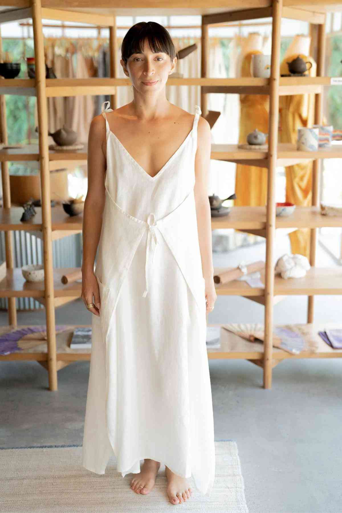 A woman stands barefoot on a rug, adorned in the Radiant Gown - Made To Order from the Myrah Penaloza collection. The sleeveless white dress, featuring a V-neck and tied straps, enhances her elegance. She is surrounded by an open wooden shelving unit filled with assorted textiles and ceramic items, evoking a warm, artisanal shop ambiance.