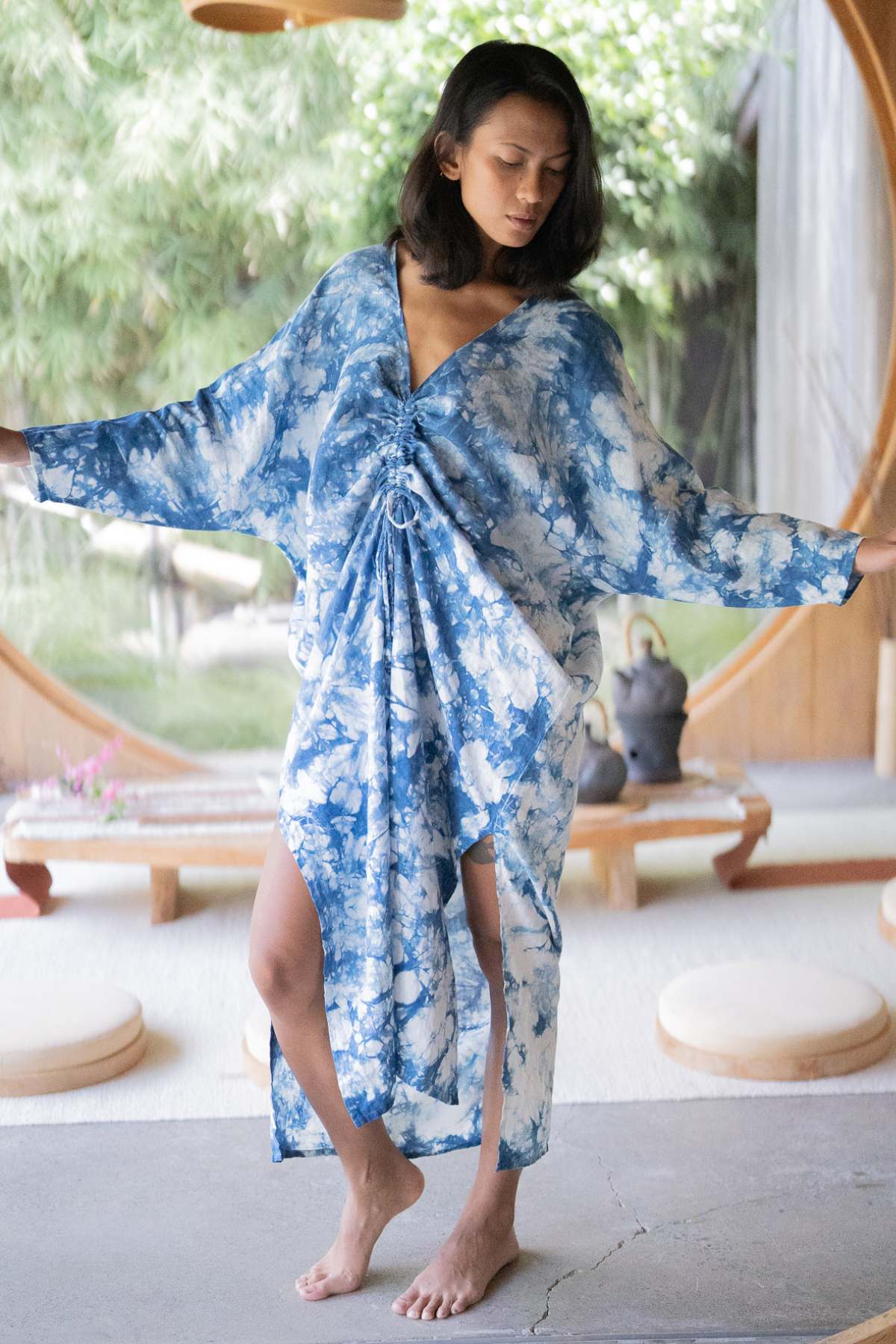 A person is standing indoors, dressed in the exclusive Indigo Virgo Rising Kaftan (One of a Kind) by Myrah Penaloza, with their arms slightly outstretched. The space is adorned with circular windows and minimalistic décor, featuring low wooden tables and cushions that mirror the serene ambiance of an Indigo Virgo Rising Kaftan.