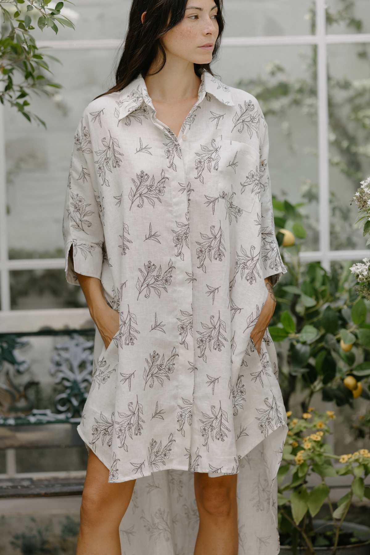 A person with long dark hair stands outdoors wearing the Linen Suka Button Down Long Top by Myrah Penaloza. This light-colored, long-sleeve top is crafted from sustainable fabric with a floral pattern. They have their hands in the pockets and are looking to the side. The background features green plants and a glass window.