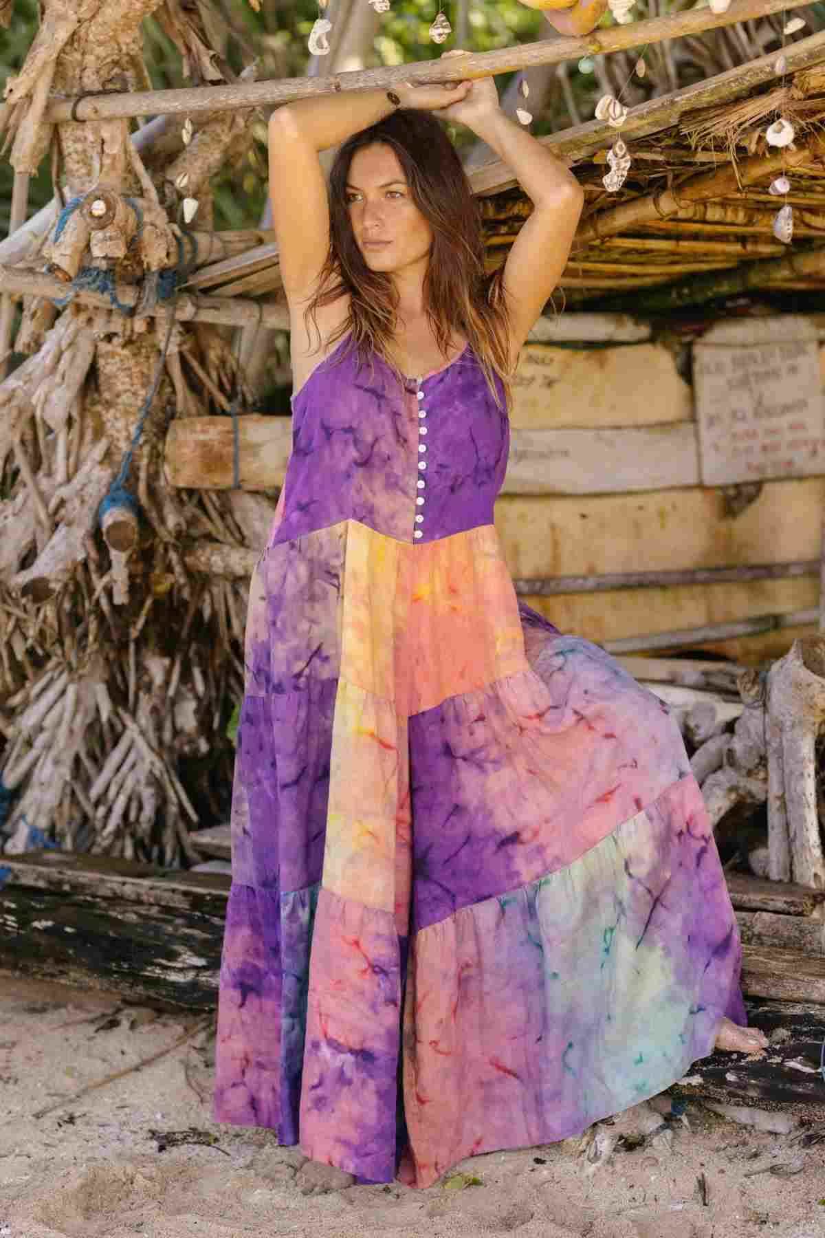 A woman wearing the La Mexicana Playsuit by Myrah Penaloza, crafted from 100% linen and made to order, stands on a sandy beach. She leans against a rustic wooden structure adorned with seashells, gazing into the distance with her arms above her head.