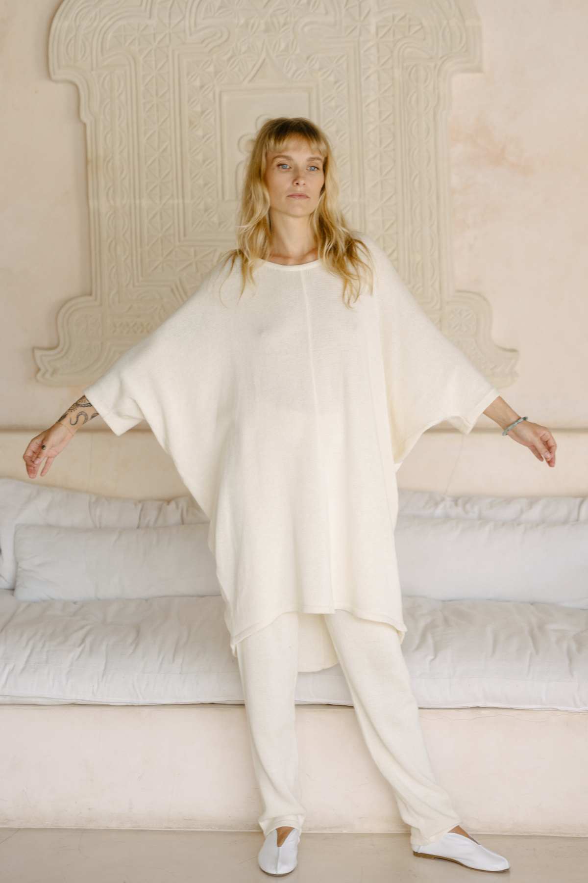 A person with long hair stands with arms slightly extended, wearing a loose, light-colored Butterfly Tunic Cotton Knit by Myrah Penaloza and matching pants. They are in front of a white, carved architectural wall detail and standing on a tiled floor. A white sofa sits behind them.