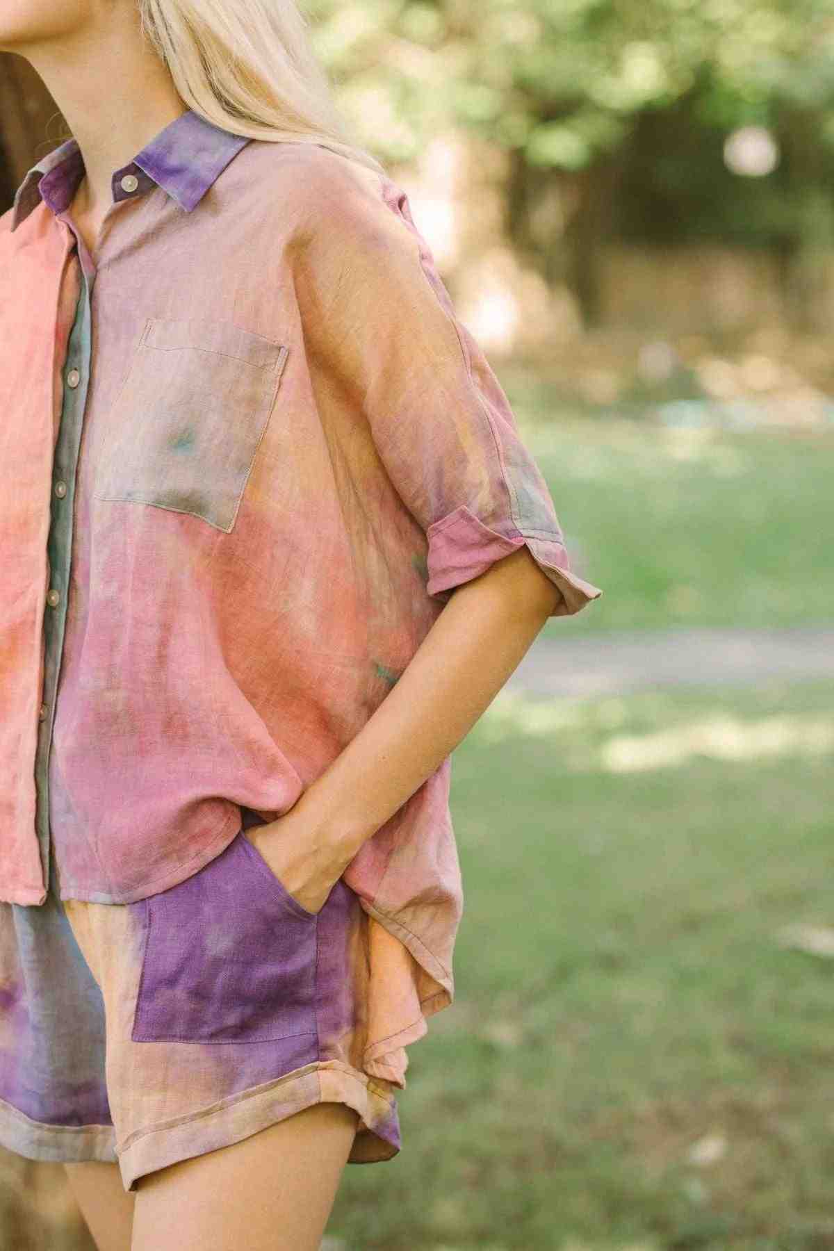 A person wearing the Suka Button Down Top from Myrah Penaloza's Rainbeau Collection, a colorful handmade tie-dye shirt crafted from ethically sourced 100% linen, stands outdoors. The shirt features rolled-up sleeves and a front pocket, with matching shorts complementing the look against a backdrop of blurred greenery.