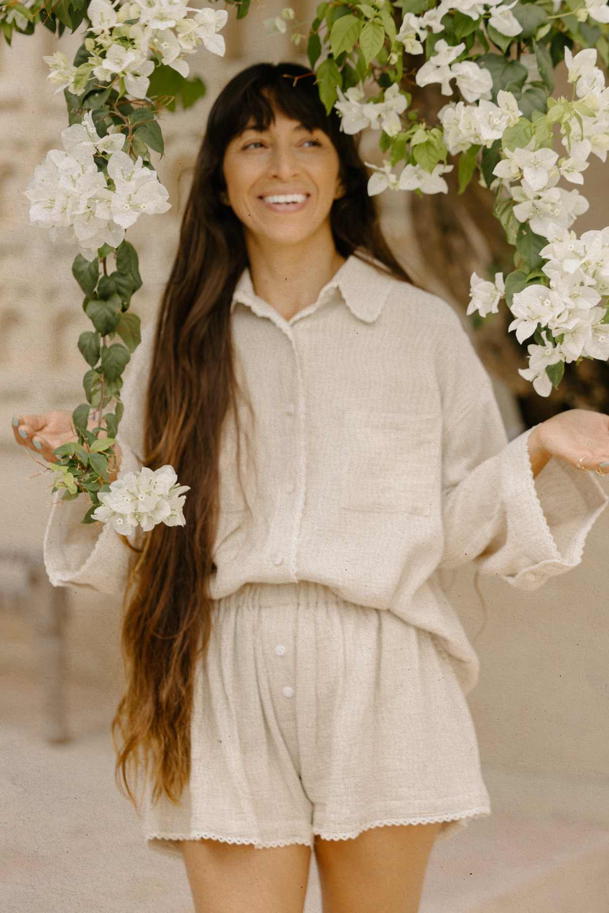 A person with long hair wearing a Nidra Button Down Set Lace Edition in beige smiles while standing next to hanging white flowers. Crafted from organic cotton by Myrah Penaloza, the outfit highlights sustainable beauty against a softly blurred background.