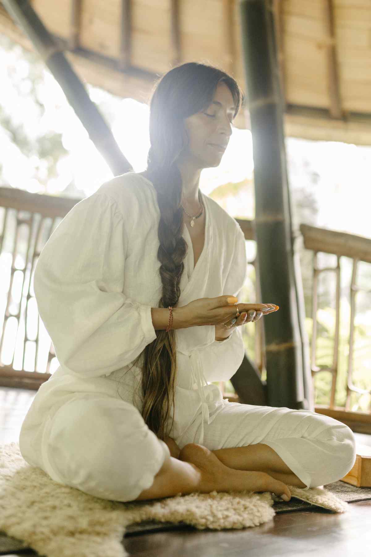 A person with long hair sits cross-legged on a mat, holding a small item with eyes closed, appearing to meditate. They are wearing a light-colored Linen Long Sleeve Dora Playsuit from Myrah Penaloza, featuring unique detailing and an adjustable waistline, in a sunlit wooden structure with a peaceful ambiance.