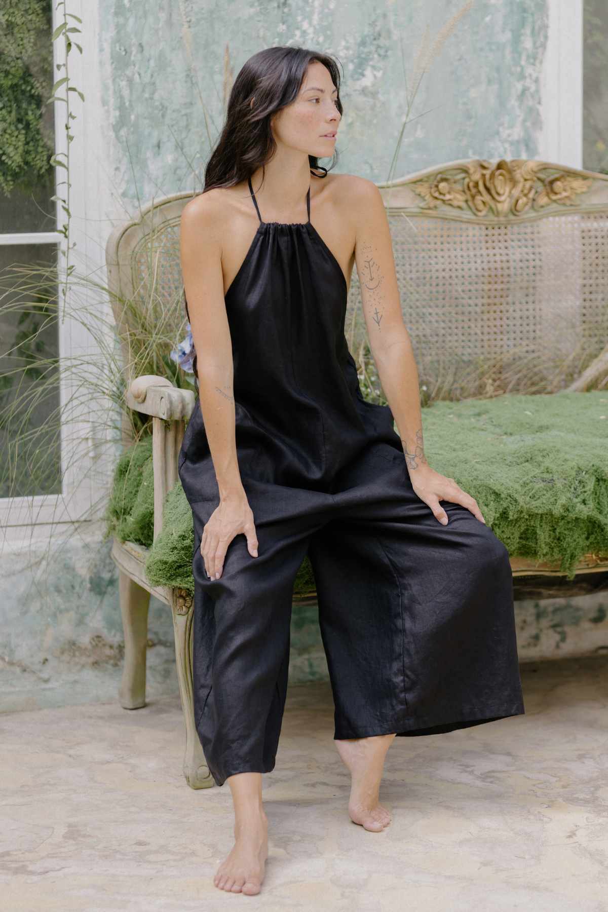 A woman with long dark hair sits barefoot on the edge of a green, vintage sofa. She is wearing the chic Vata Hari Playsuit by Myrah Penaloza, which features a stylish black halter-style neckline, and has tattoos on her right arm. The background includes a window against a worn, green wall.