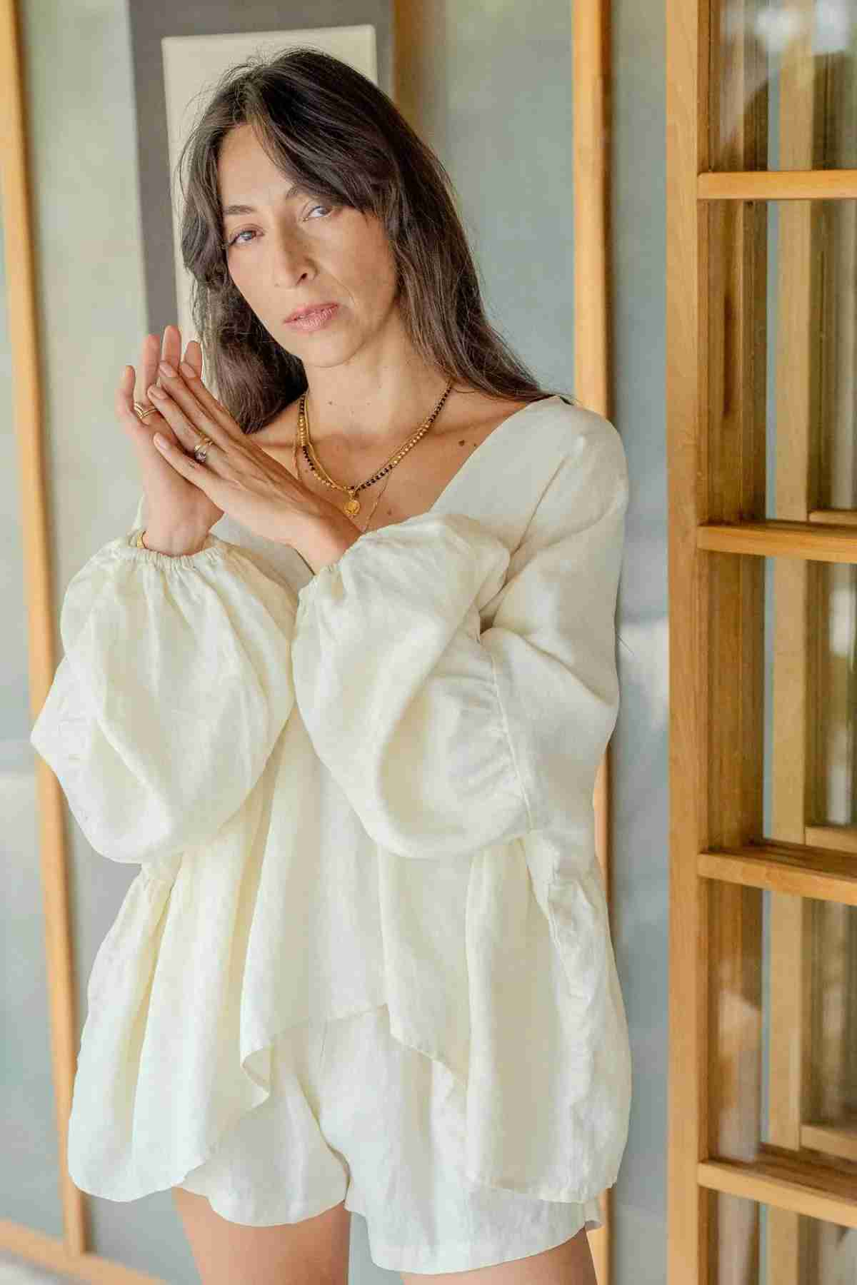 A woman with long dark hair stands in a brightly lit room, wearing a loose-fitting Eva Linen Top by Myrah Penaloza with flowering sleeves and white shorts. She has her hands clasped together near her face and is gazing directly at the camera.