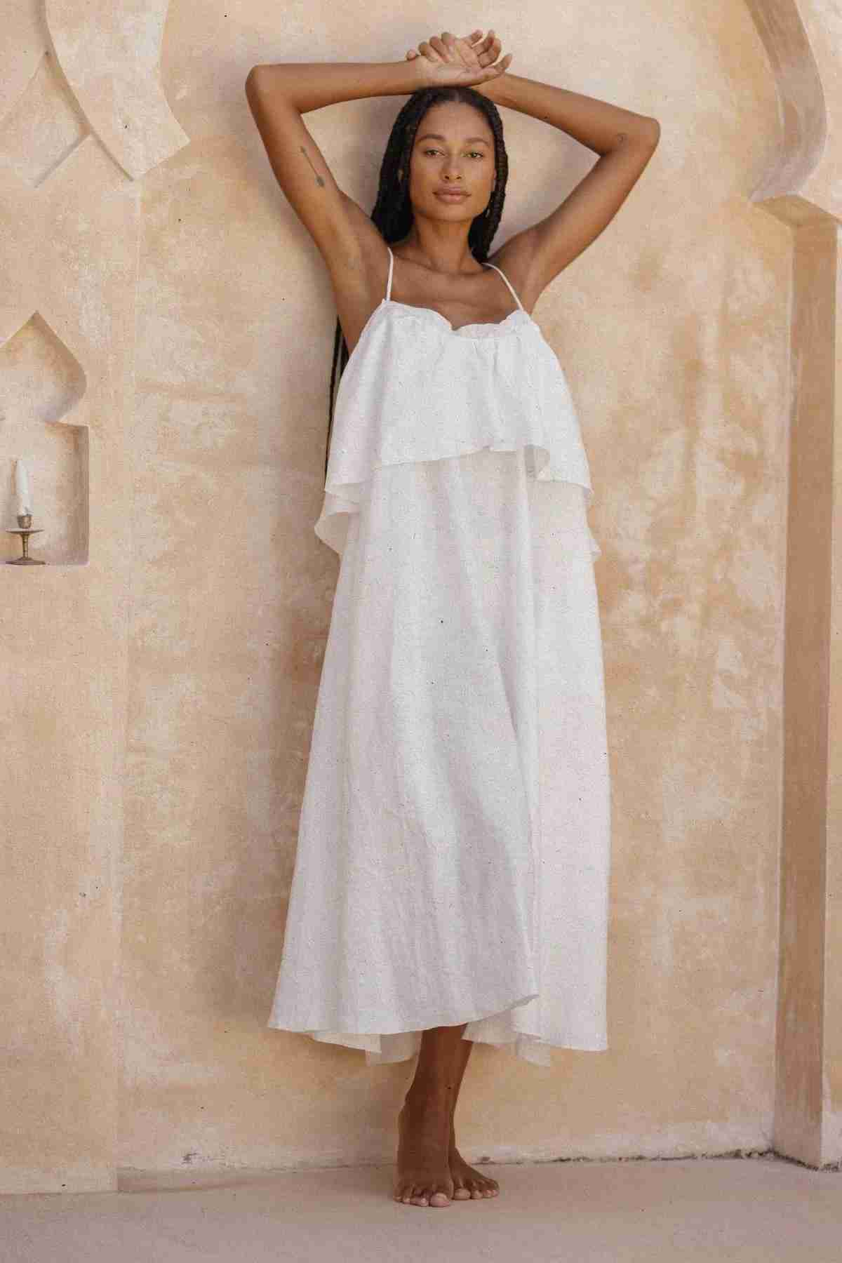 A woman with long braided hair stands barefoot against a textured beige wall. She wears the Linen Muse Gown by Myrah Penaloza, a flowing white dress with adjustable straps and a low back. She holds her arms above her head, touching the wall. The background features arched details and a small alcove with a candle.