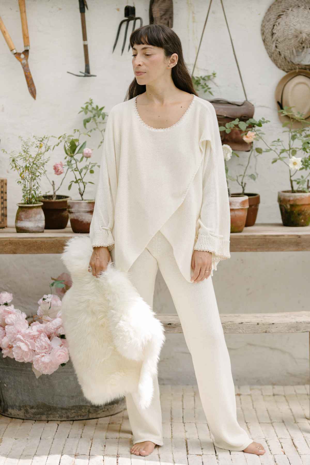 A woman stands in a rustic garden room, dressed in a cream Honey Knit Sweater by Myrah Penaloza, holding a fluffy white blanket. Around her are potted plants, gardening tools on the wall, and a basket of pink flowers on the floor, creating a serene and cozy atmosphere.