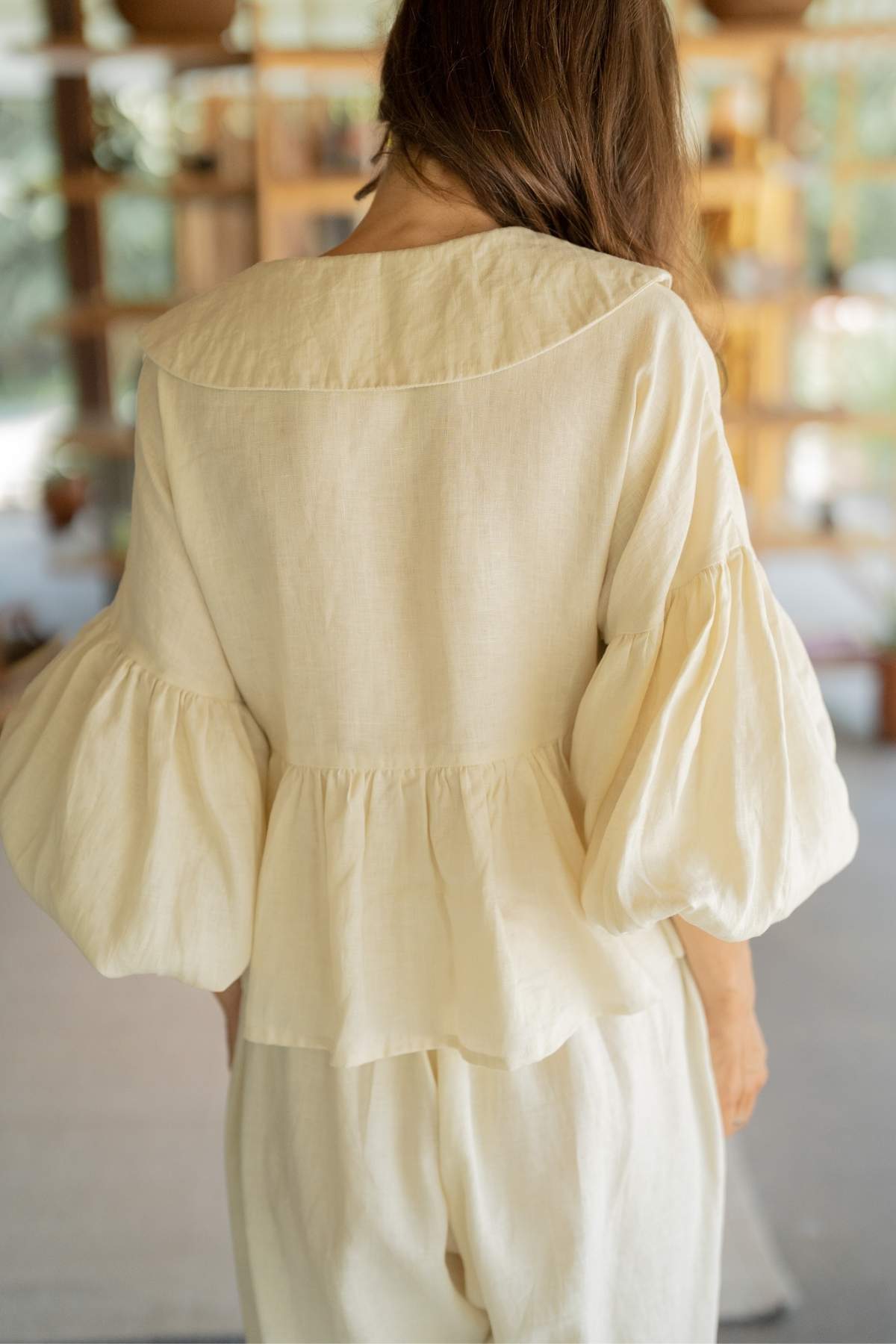 A person with long brown hair is seen from behind, wearing a light beige, long-sleeve, loose-fitting blouse with puffed sleeves. The Magdalena Wrap Top Set by Myrah Penaloza features a gathered waist detail and is paired with matching Donna Pants. The background is softly blurred.