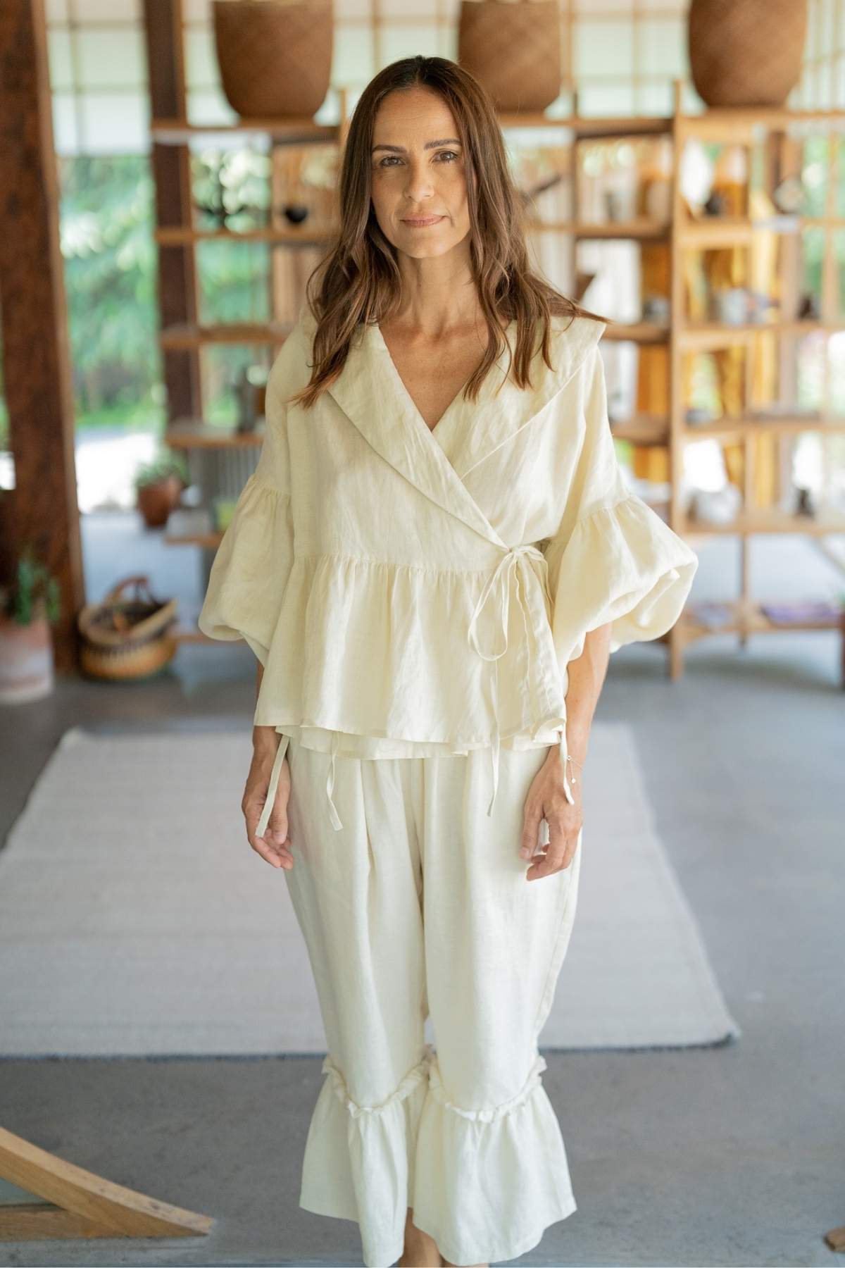 A woman stands indoors wearing the chic Magdalena Wrap Top Set with Donna Pants by Myrah Penaloza, featuring a cream-colored, loose-fitting wrap top with puffy sleeves and matching wide-legged pants. She is in a room adorned with wooden shelves that display various decorative items and plants, as sunlight filters through the windows in the background.