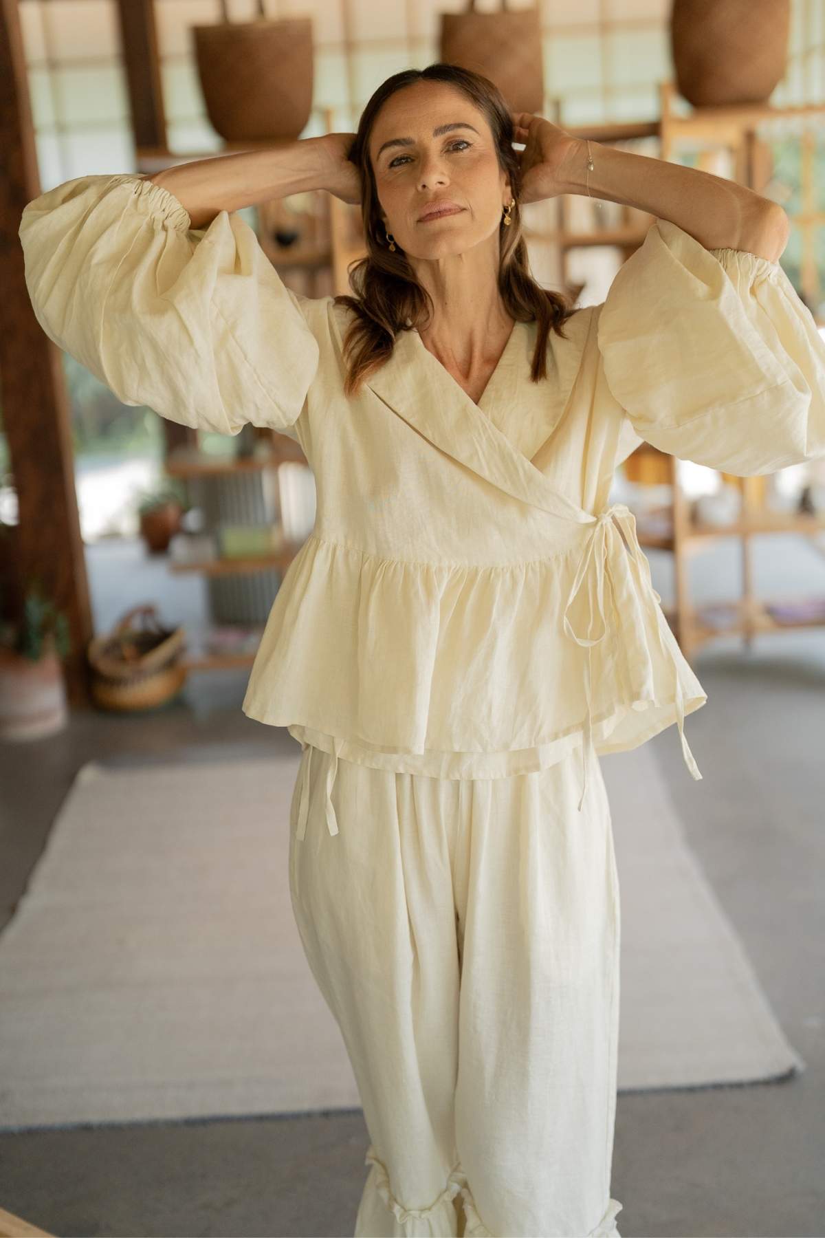 A woman stands indoors wearing a light, flowing Magdalena Wrap Top Set with Donna Pants from Myrah Penaloza. The wrap top has billowy sleeves that add to her chic appearance. She has long hair and is holding it back with both hands. Wooden shelves and various decorative items in the background enhance her effortlessly stylish look.