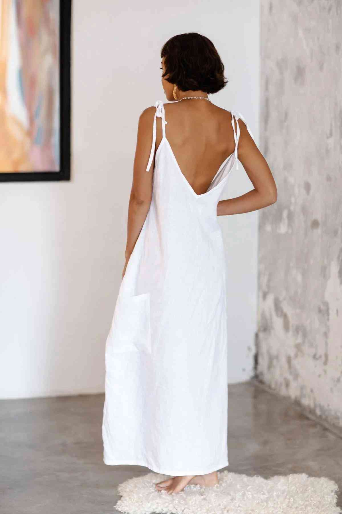 A person with short, dark hair stands barefoot on a white textured rug in a minimalist room. They are wearing the long, sleeveless Radiant Gown - Made To Order from Myrah Penaloza's healing linen collection, crafted from 100% Linen, featuring a low back and shoulder tie straps. They gaze at the wall adorned with modern abstract artwork.