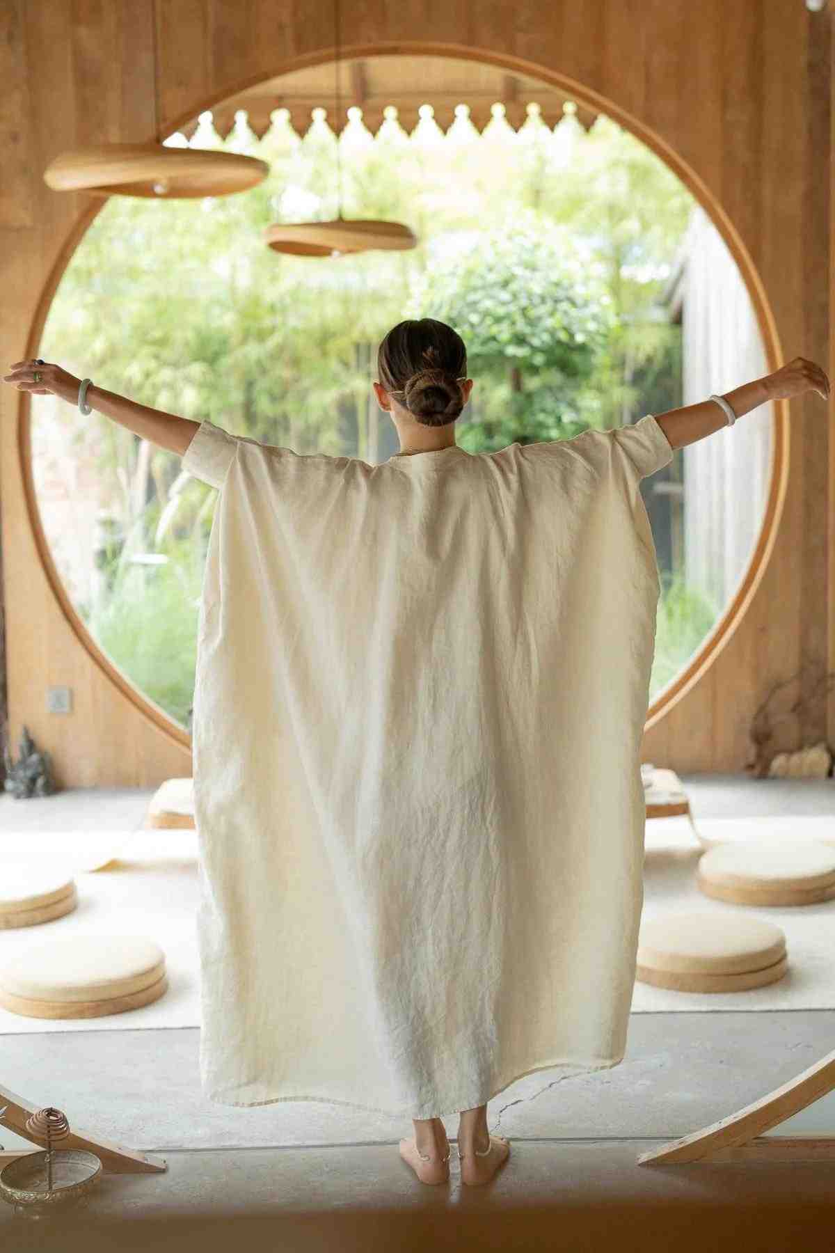 A person with a neatly tied bun stands with arms outstretched, facing away from the camera. They are wearing a loose, white La Majia Kaftan by Myrah Penaloza in a wooden room with a large circular window. Greenery is visible through the window, and there are circular cushions on the floor.