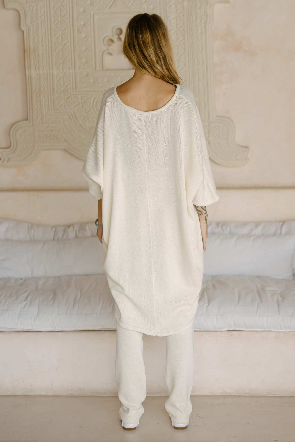 A person with long, light brown hair is standing and facing away from the camera, dressed in loose, cream-colored clothing. They are wearing the Butterfly Tunic Cotton Knit by Myrah Penaloza and matching pants made from sustainable fabric. The backdrop features a neutral-toned wall with decorative detailing and a white cushioned bench.