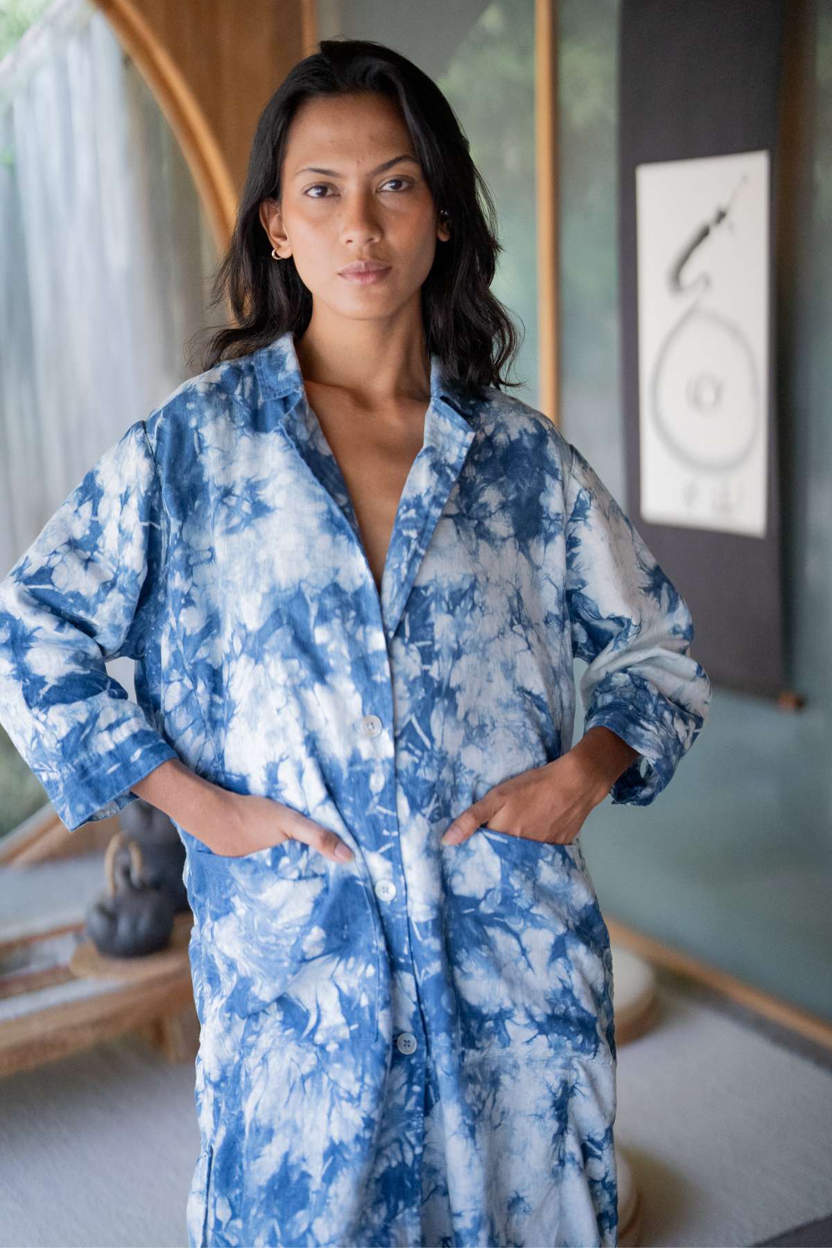 A woman stands indoors wearing the Rubi Linen Jacket Indigo by Myrah Penaloza, available during Black Friday. She has her hands in the jacket's pockets. The background features a wooden arch, a small statue, and a framed black and white artwork.