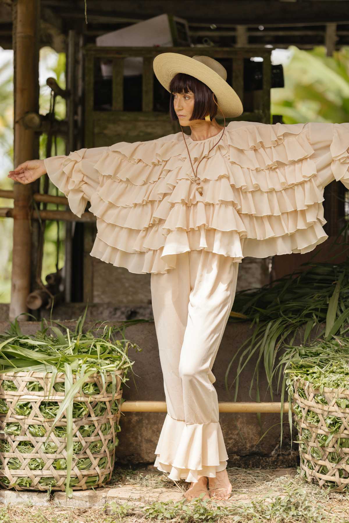 A woman stands in a garden wearing the Anusara Bloom - Made To Order by Myrah Penaloza, a flowing, layered cream-colored outfit with ruffled sleeves and pants. Crafted from chiffon sheer silk, this limited edition ensemble features a wide-brimmed hat. She poses with her arms slightly outstretched beside two large woven baskets filled with green plants.