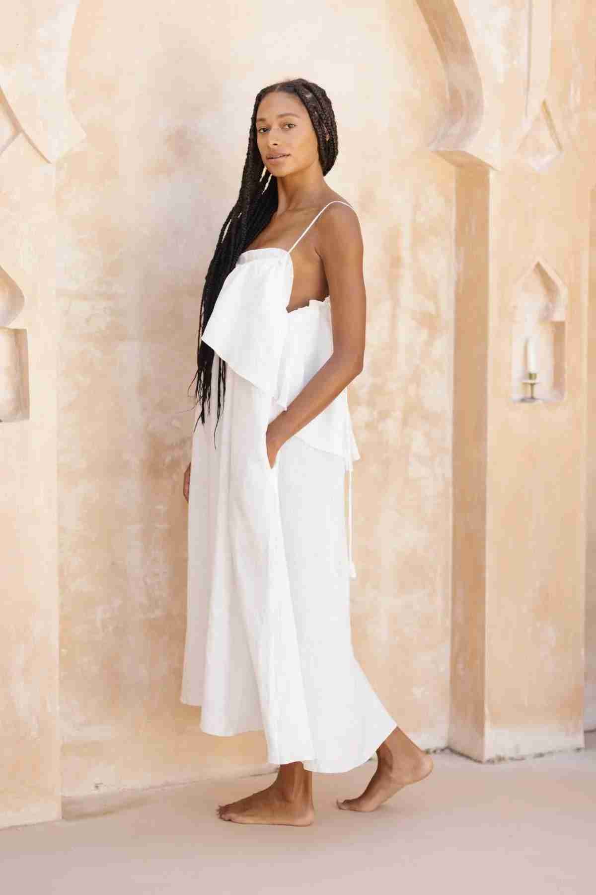 A woman with long braided hair is standing barefoot in a softly lit, archway-adorned room. She is wearing the Linen Muse Gown by Myrah Penaloza—a flowing, white sleeveless, stone washed linen dress with adjustable straps—and looking confidently at the camera. The background consists of warm-toned walls with small, candlelit niches.