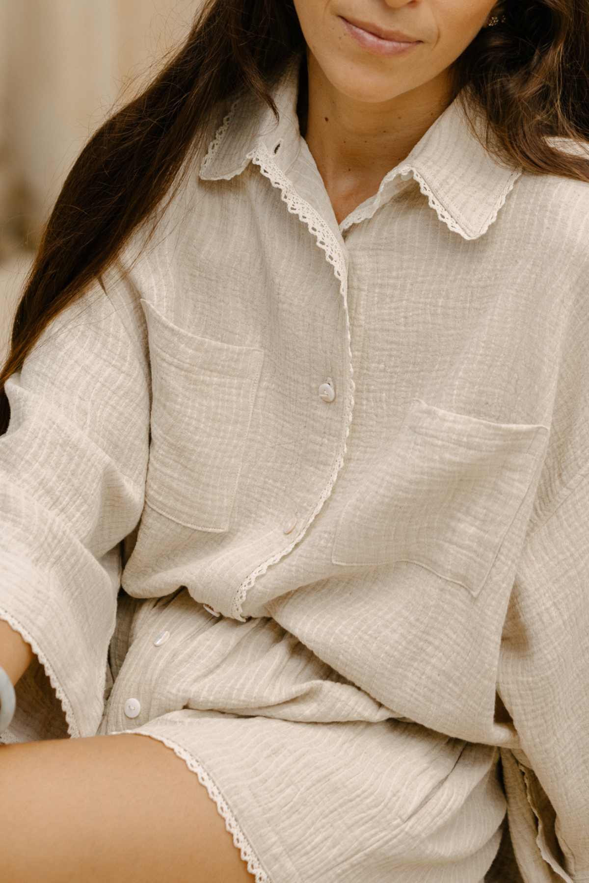 A woman is wearing the Nidra Button Down Set Lace Edition from Myrah Penaloza in cream, made from organic cotton. The ensemble includes lace-trimmed shorts and displays a relaxed, textured fabric with two chest pockets and white buttons. Her long brown hair flows freely, partially obscuring her face, where she smiles slightly.