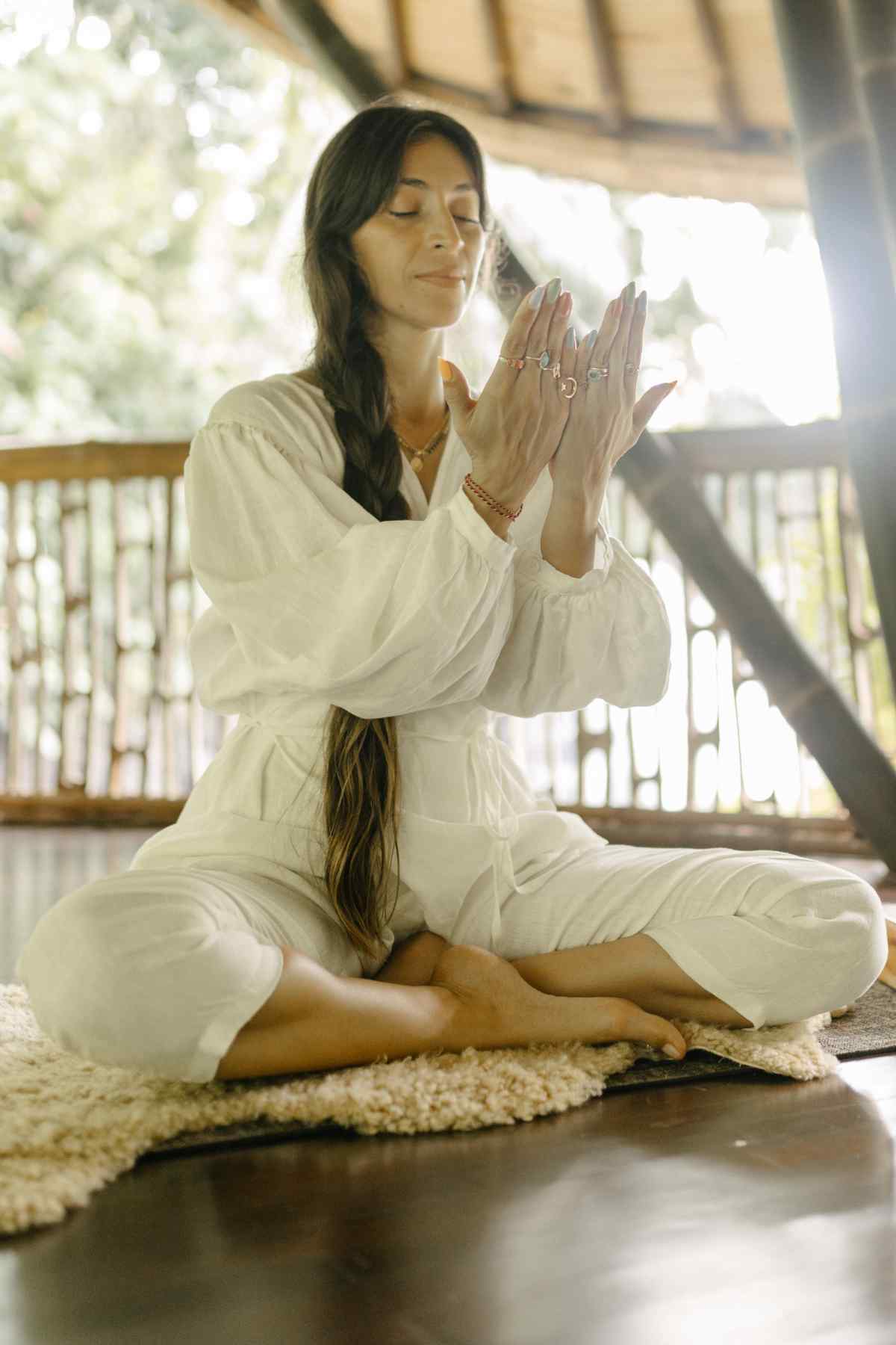 A person with long hair sits cross-legged on a rug indoors, wearing a white 100% Linen Long Sleeve Dora Playsuit by Myrah Penaloza, featuring an adjustable waistline. They have their eyes closed and hands together in front of their face as if meditating or practicing yoga. Sunlight filters through the bamboo structure in the background.