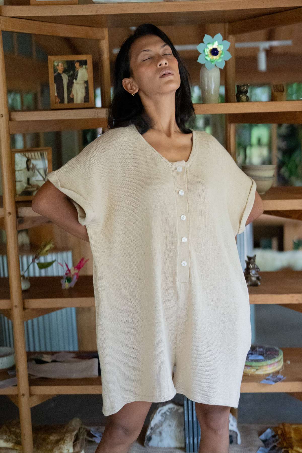 A woman stands confidently with her eyes closed and hands on her hips, wearing the Henley Knitted Playsuit - BF by Myrah Penaloza. She is positioned in front of wooden shelves adorned with various decorations and photos.