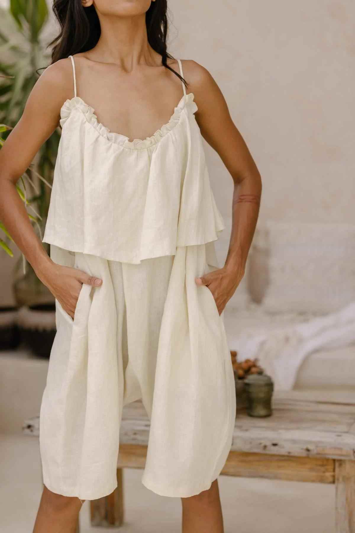 A person stands indoors wearing a loose, off-white, flowing Muse Linen Playsuit by Myrah Penaloza, made from stone-washed linen with thin, adjustable straps. The playsuit has a layered design and features a low back cut. Their hands rest in the pockets of the playsuit. The background includes a wooden table, some plants, and a neutral-colored wall.