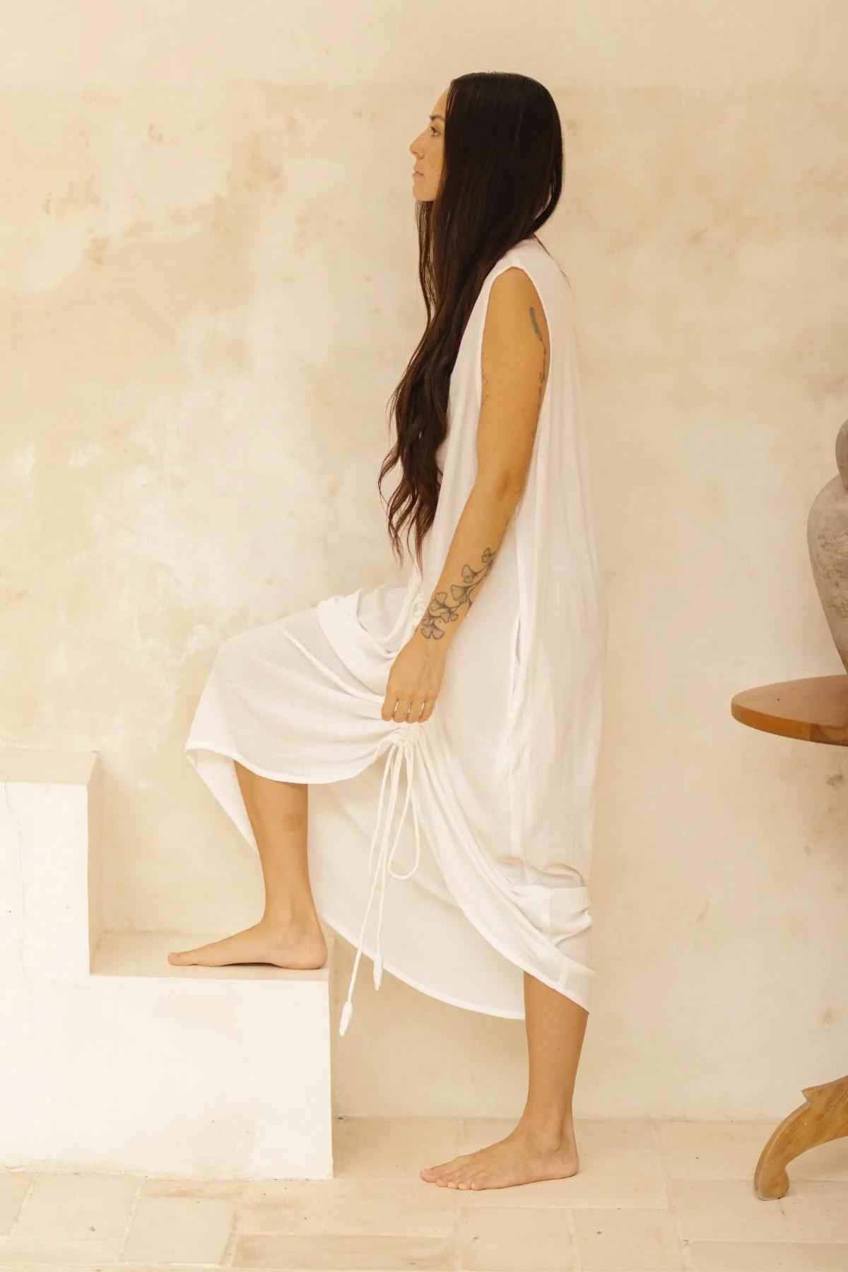 A person with long, dark hair and visible tattoos is dressed in the sleeveless Myrah Penaloza Virgo Luna Dress, standing barefoot on steps against a minimalist, light-colored background. They appear to be mid-movement, looking ahead while holding part of their dress.