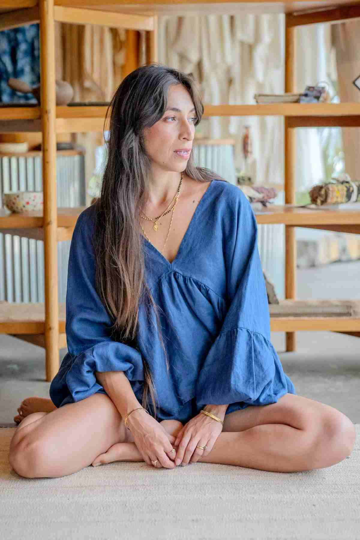 A woman with long dark hair sits cross-legged on the floor, wearing a flowing blue Eva Linen Top from Myrah Penaloza, featuring flowering sleeves. She is looking to her left, and behind her are wooden shelves filled with various items. The setting appears relaxed and natural.