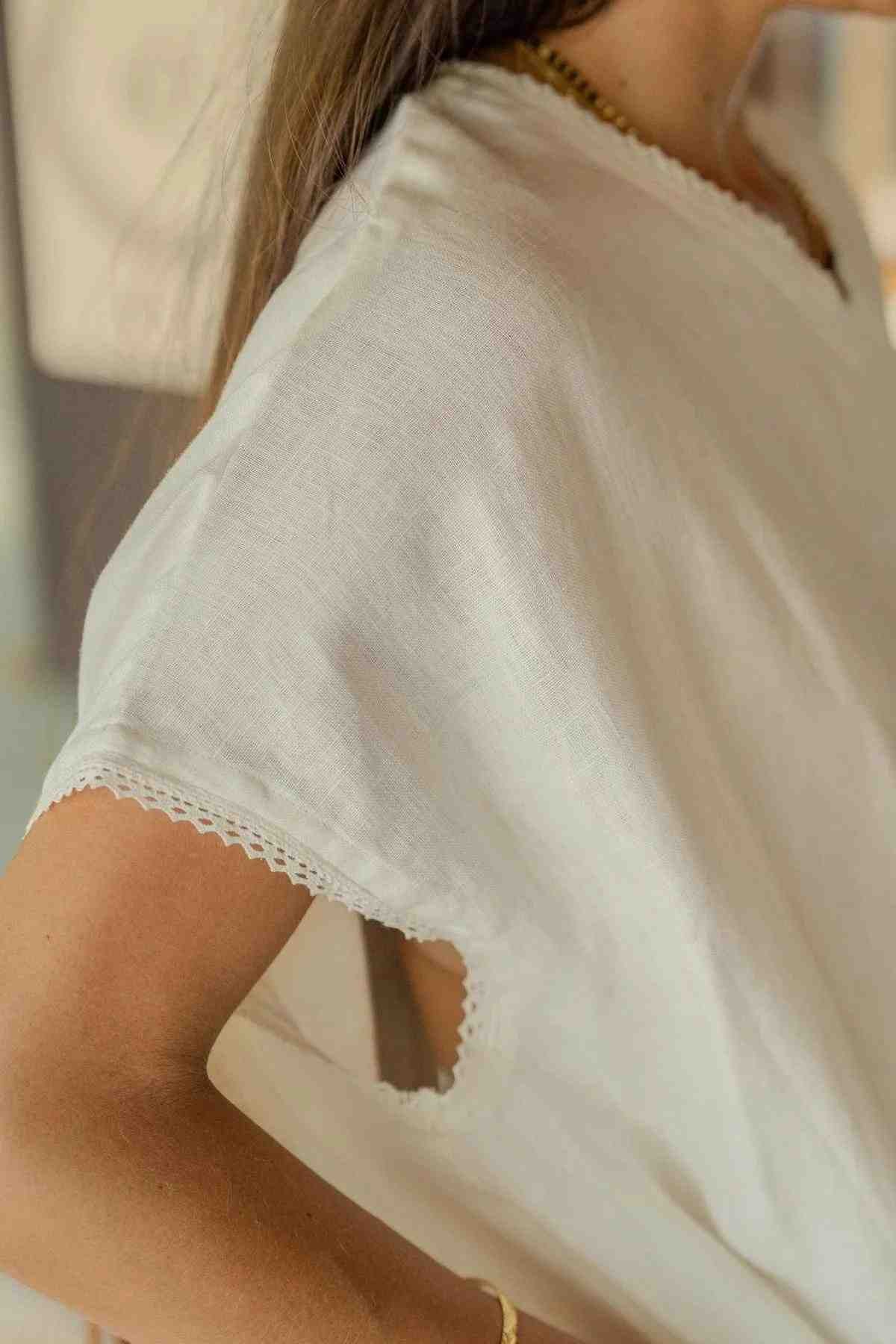 Close-up of a person wearing the Ruby Lace Tunic by Myrah Penaloza—a white, loose-fitting, short-sleeve lace tunic with a lace-trimmed V neckline and sleeves. The person's arm is bent at the elbow, hand resting on their hip. A light-colored background and partial view of a necklace are visible.