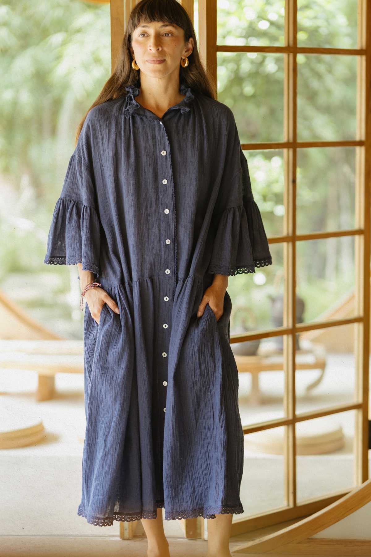 A woman stands indoors wearing an elegant Myrah Penaloza Juliette Linen Gown, complete with ruffled sleeves. Her long hair flows down as she places her hands in the gown's pockets. The backdrop showcases large windows that provide a view of the lush greenery outside.