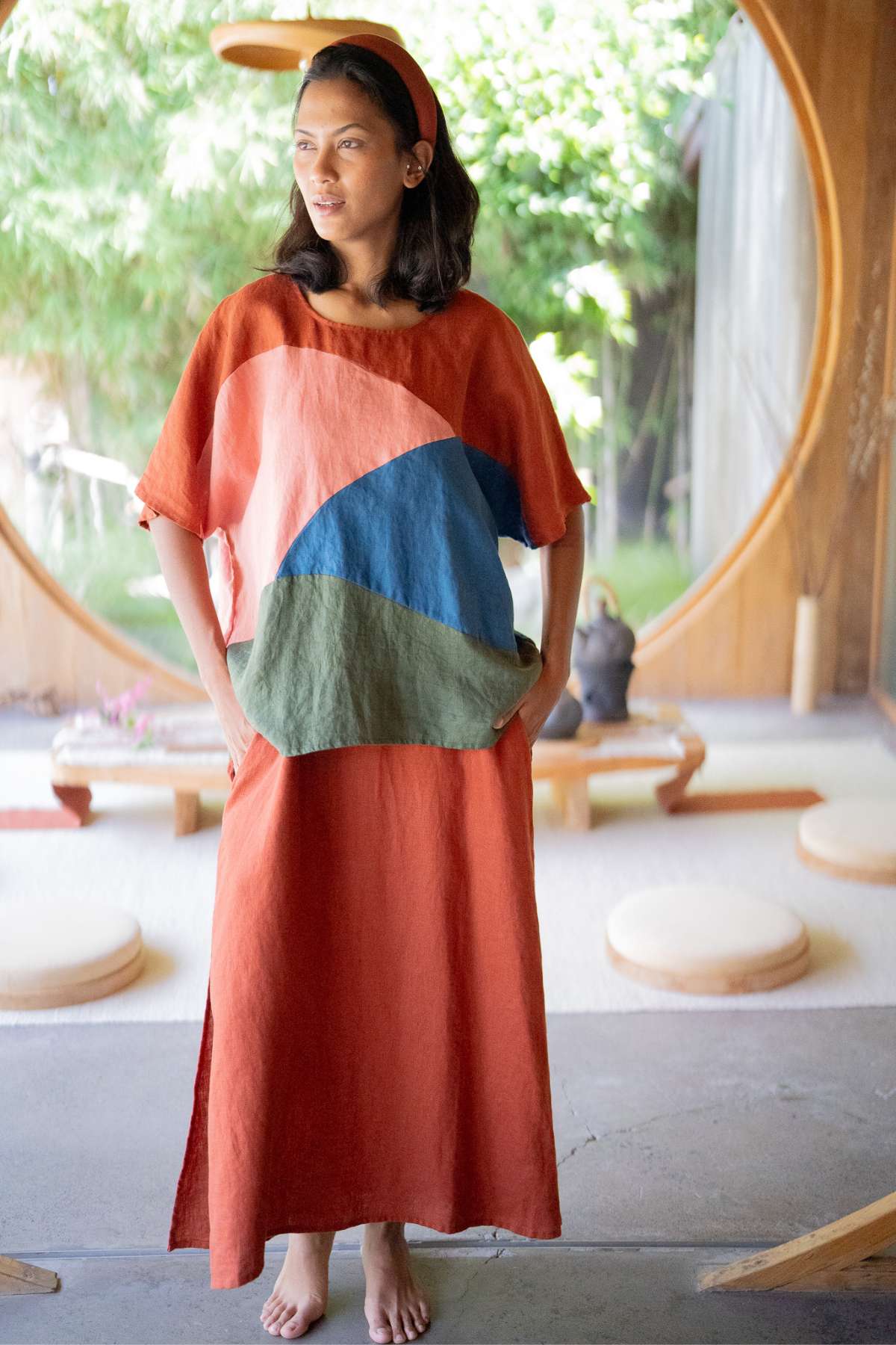 A woman stands barefoot in a sunlit room with a large circular window. She is wearing the Uluwatu Top by Myrah Penaloza, featuring a colorful pattern of abstract blue and green shapes in rust tones, made from sustainable linen. Her dark hair is styled with a headband, and she casually places her hands in the top's pockets.