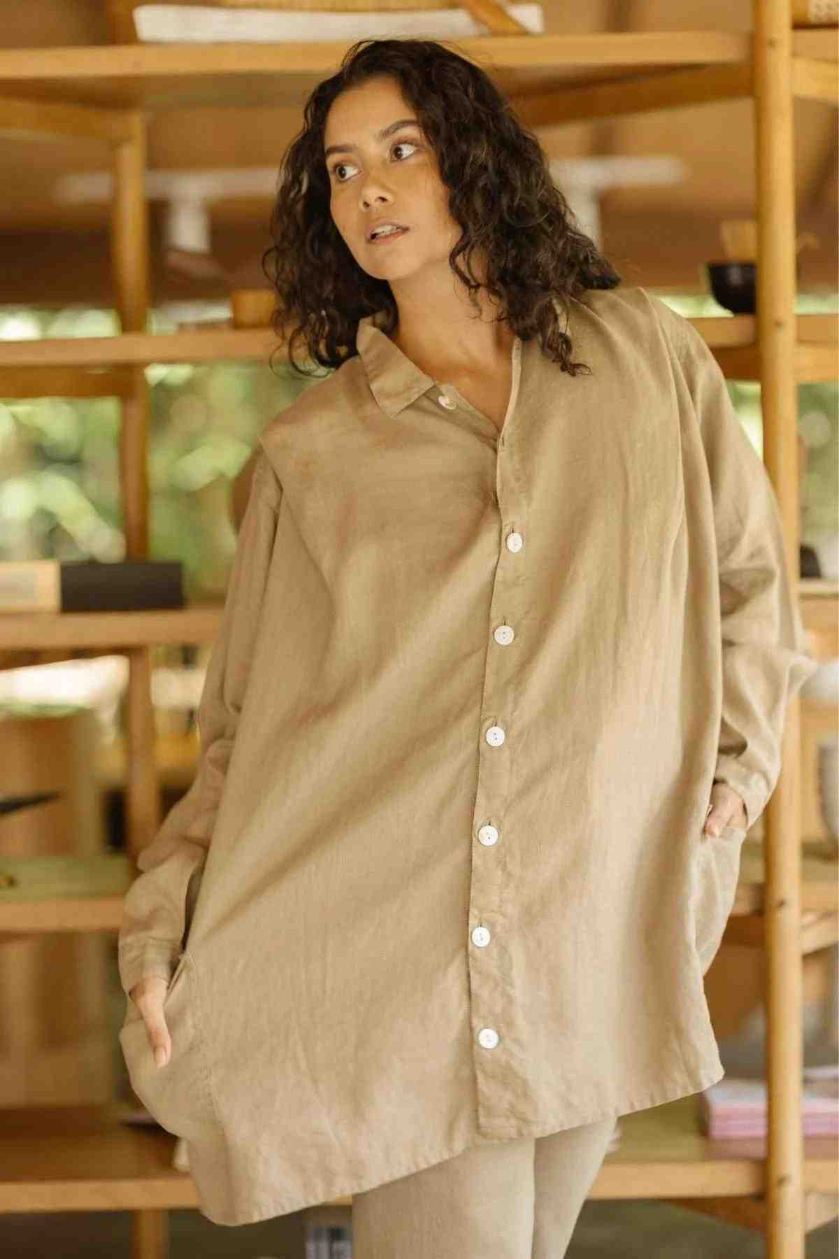 A woman with curly hair wearing an oversized beige button-up shirt poses indoors, showcasing her eclectic fashion sense. In the background, a wooden shelf filled with various items forms a cozy setting. She completes her look with the eye-catching Tender Jacket (100% Linen) by Myrah Penaloza, draped over one shoulder in a deep royal blue hue.