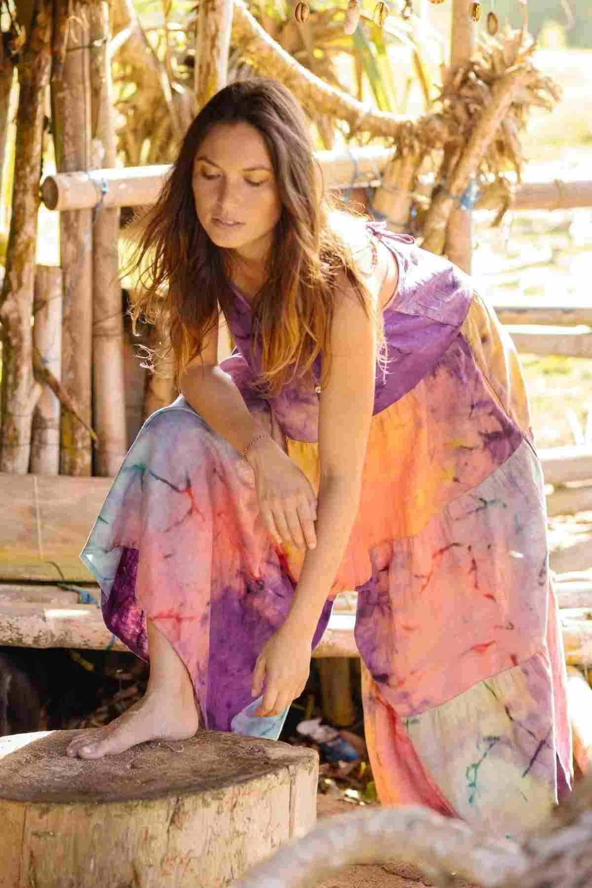 A woman models the La Mexicana Playsuit by Myrah Penaloza, crafted from 100% linen and made to order. She leans forward with one foot on a wooden stump, surrounded by a rustic structure of wooden beams and ropes. The playsuit's adjustable straps rest gently on her shoulders as sunlight casts soft shadows around them.