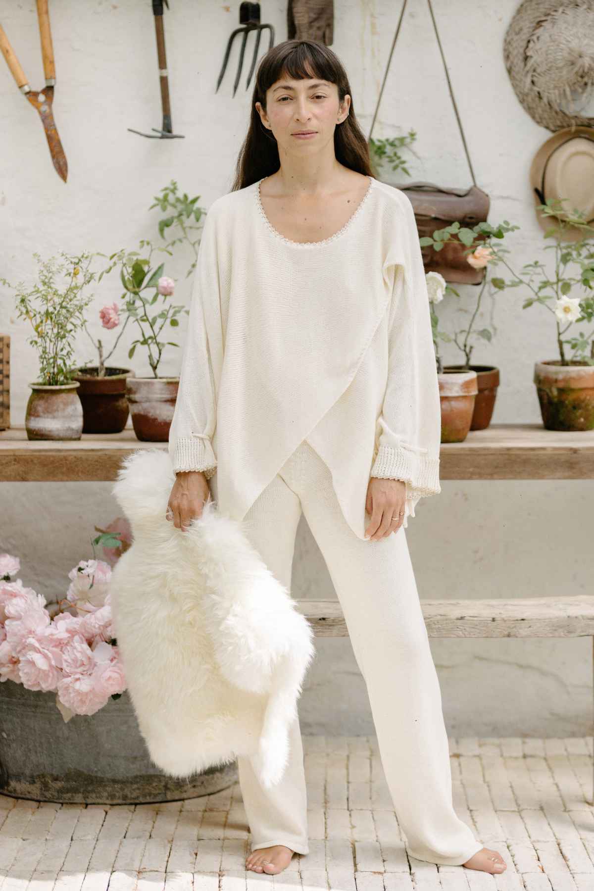 A woman stands in a rustic setting, wearing the Honey Knit Sweater from Myrah Penaloza paired with coordinating pants. She holds a fluffy, white throw in her right hand. Behind her are potted plants and hanging garden tools on a white wall, with more plants and pink flowers adorning the surroundings.