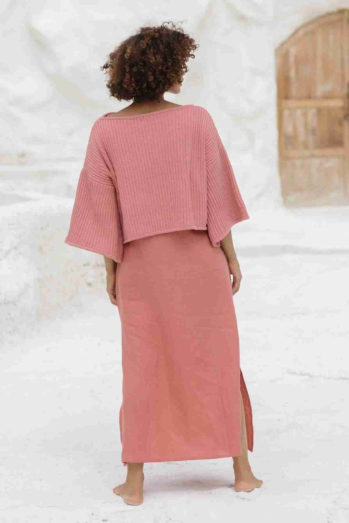 A person with curly hair is standing barefoot on a white surface, facing away from the camera. They are wearing the oversized, pink Spirit Top Sweater Knit by Myrah Penaloza, featuring wide sleeves and made of luxurious cotton fabric. They are also dressed in a matching ankle-length skirt with a slit on one side. A wooden door is visible in the background.