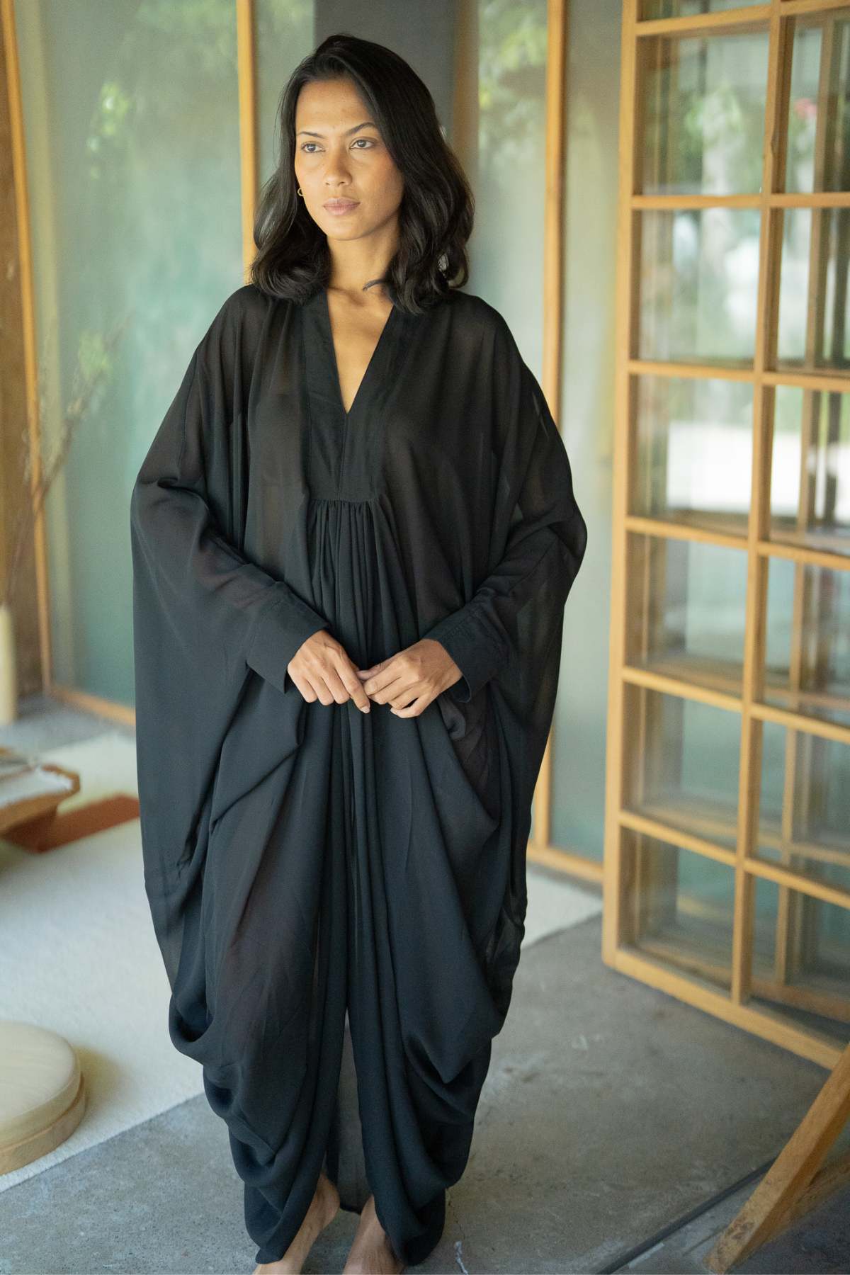 Wearing the opulent Myrah Penaloza Leo Kaftan Chiffon Sheer, a person stands in a tranquil room adorned with wooden accents and gentle lighting, gazing thoughtfully into the distance. The ambiance exudes calmness and peace.