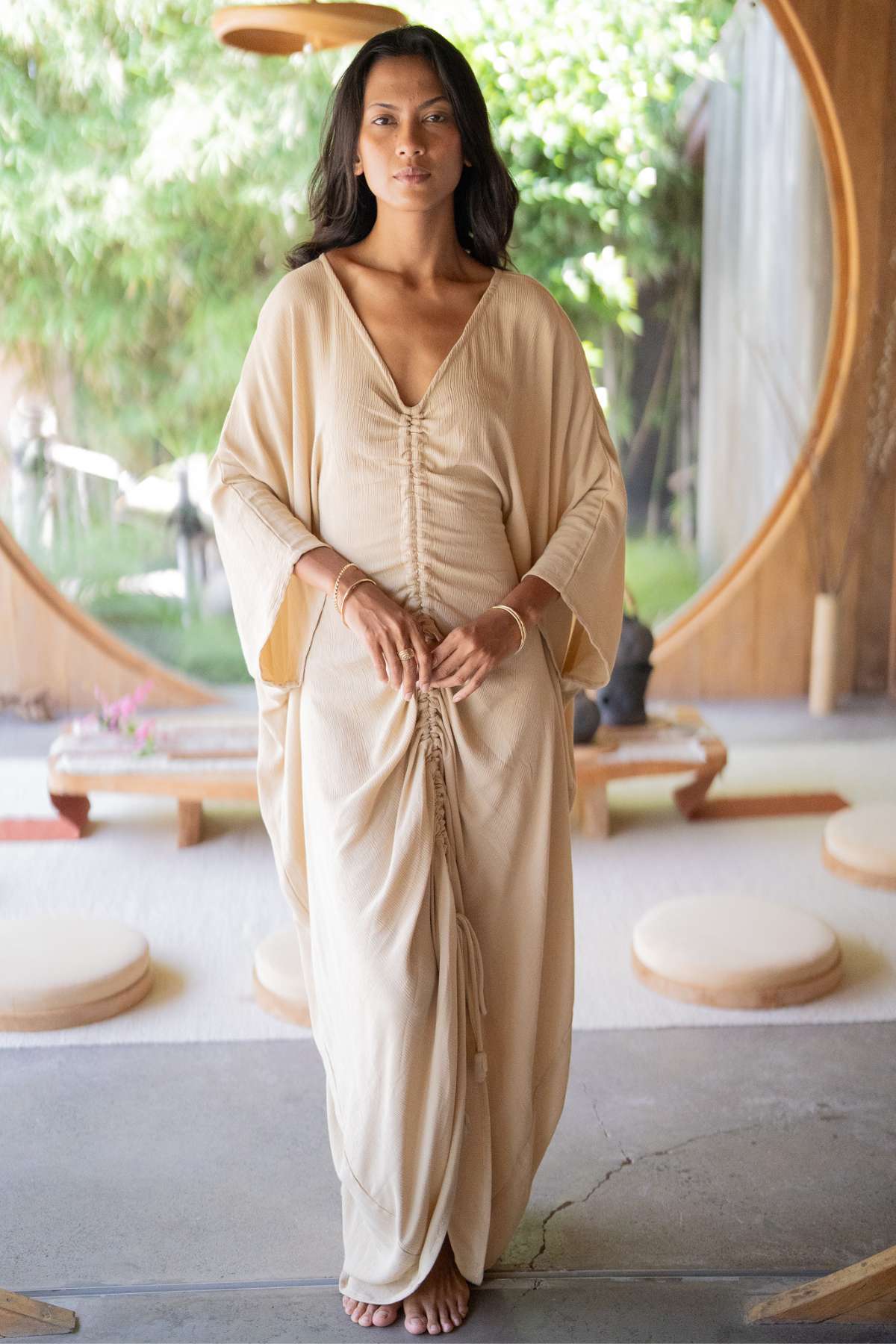 In a sunlit room, an individual wears a long, beige Virgo Moon dress by Myrah Penaloza, featuring gathered details. Circular windows and minimalistic wooden furniture in the background create a serene, natural atmosphere with a hint of ceremonial whimsy.