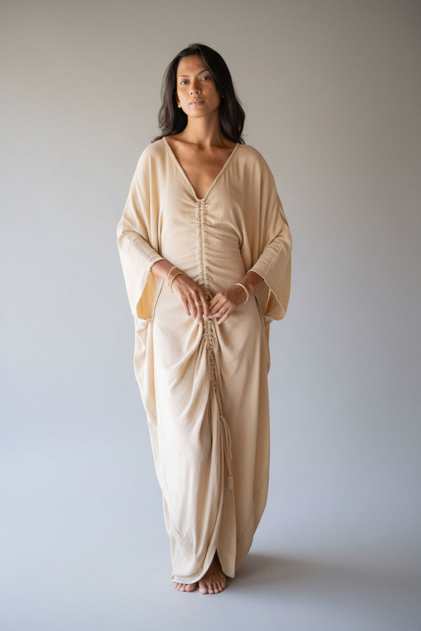 In a sunlit room, an individual wears a long, beige Virgo Moon dress by Myrah Penaloza, featuring gathered details. Circular windows and minimalistic wooden furniture in the background create a serene, natural atmosphere with a hint of ceremonial whimsy.