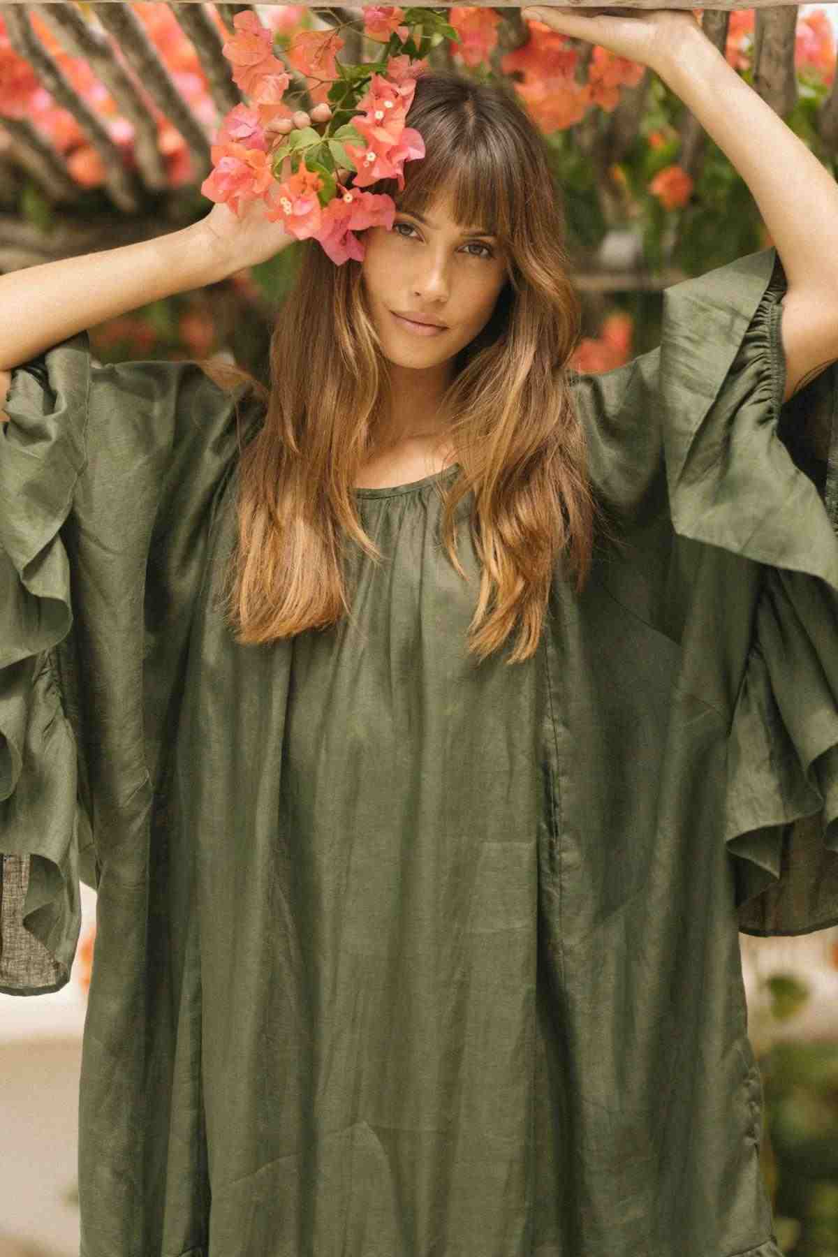 A person in a Brida Gown Long by Myrah Penaloza stands beneath flowering branches, holding some orange-pink blossoms near their head. The scene is tranquil and natural, with soft lighting enhancing the relaxed atmosphere.