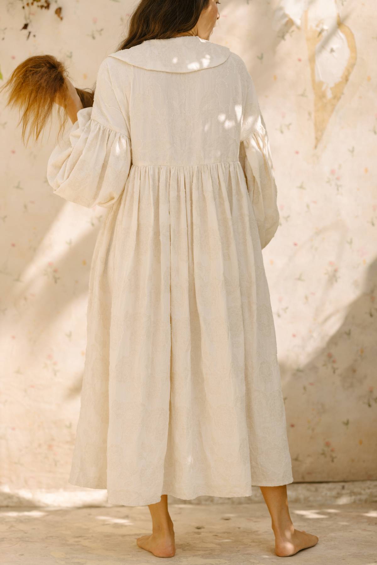 A woman in the Blooming Magdalena Dress by Myrah Penaloza stands barefoot with her back to the camera, holding a tall grass stalk. The long, flowing white dress features puffed sleeves and subtle textured patterns. Dappled light creates soft shadows on the wall and her dress.