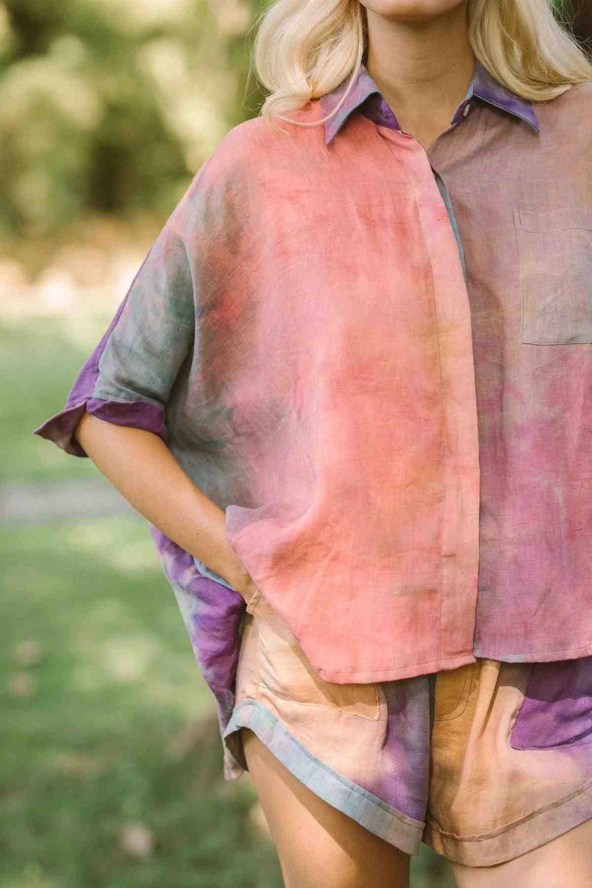 A person is standing outdoors wearing the Suka Button Down Top from Myrah Penaloza's Rainbeau Collection, made of 100% sustainable linen. The outfit features matching shorts and showcases a blend of pastel pink, purple, and blue-green shades against a blurred green landscape.