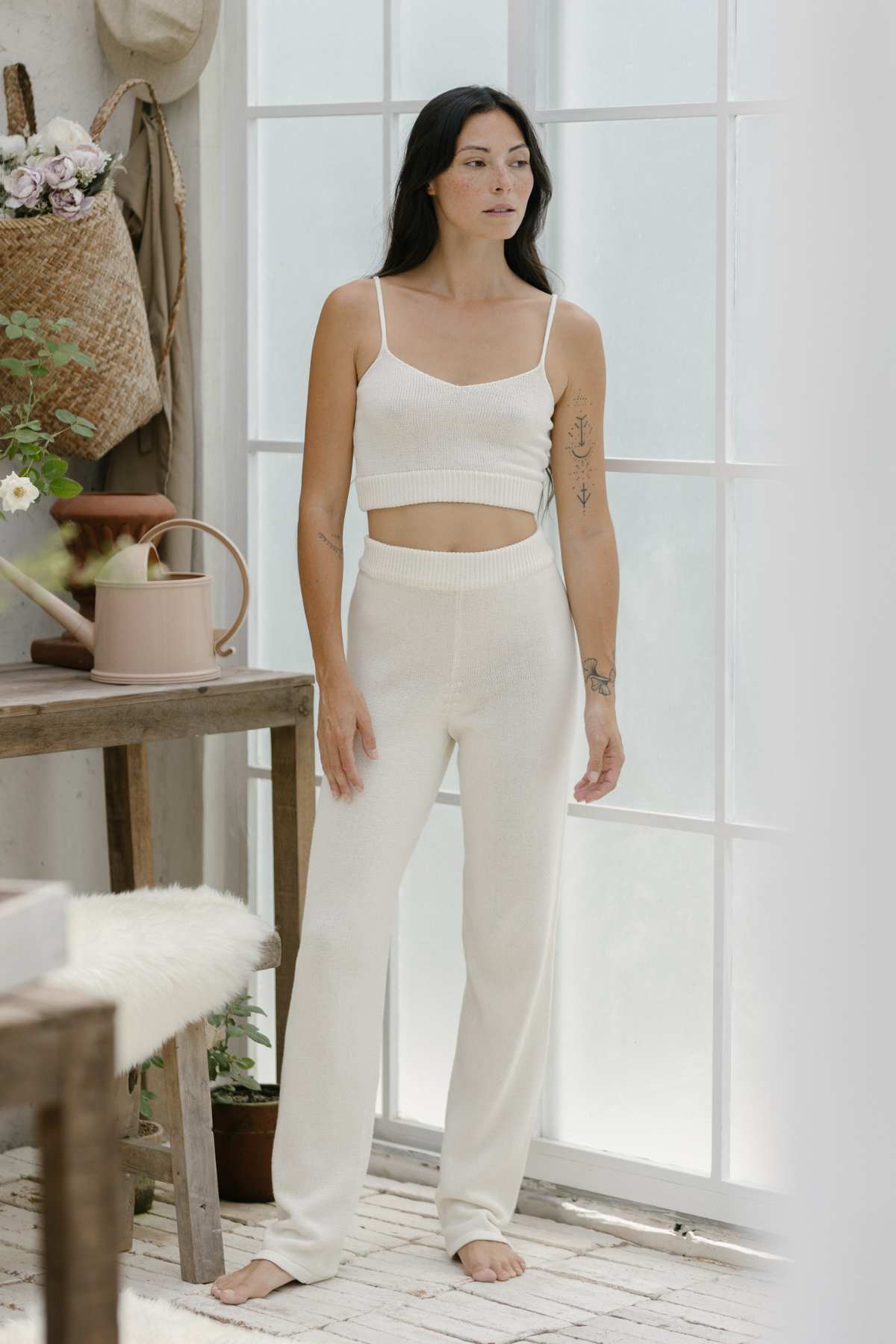 A person with long dark hair stands indoors wearing a light-colored sleeveless top and matching pants from the Honey Knit Sets by Myrah Penaloza. Tattoos adorn their arm as they stand near a wooden table with a pink watering can and flowers. Soft light filters through the large window, highlighting the scene.