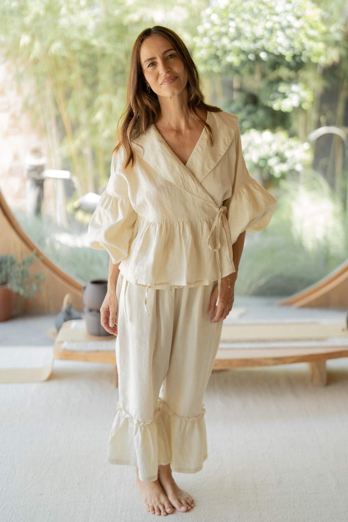 A woman with long hair stands barefoot indoors, exuding effortless chic in the Magdalena Wrap Top Set with Donna Pants by Myrah Penaloza. She is wearing a light-colored, loose-fitting wrap top paired with flared pants. The background showcases lush greenery through large circular windows, and the room reflects a serene, minimalist aesthetic.