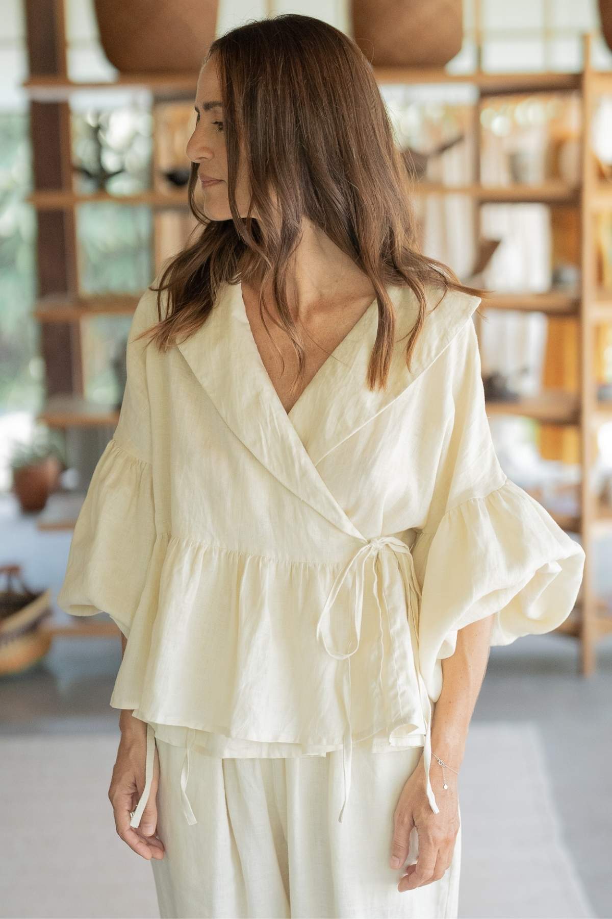 A woman with long brown hair is wearing a chic ensemble from Myrah Penaloza—a beige Magdalena Wrap Top Set with puffed, three-quarter-length sleeves made of Moonlight linen fabric, paired with Donna Pants. The setting appears to be a well-lit, modern room with shelves in the background. She is looking to her left and has a relaxed expression.