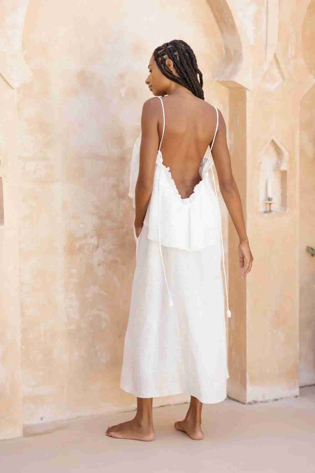 A woman with braided hair stands barefoot, facing away from the camera. She is wearing the Linen Muse Gown by Myrah Penaloza, a flowing, stone-washed linen dress with adjustable straps that reveal a low back. The background features a beige wall with architectural details.